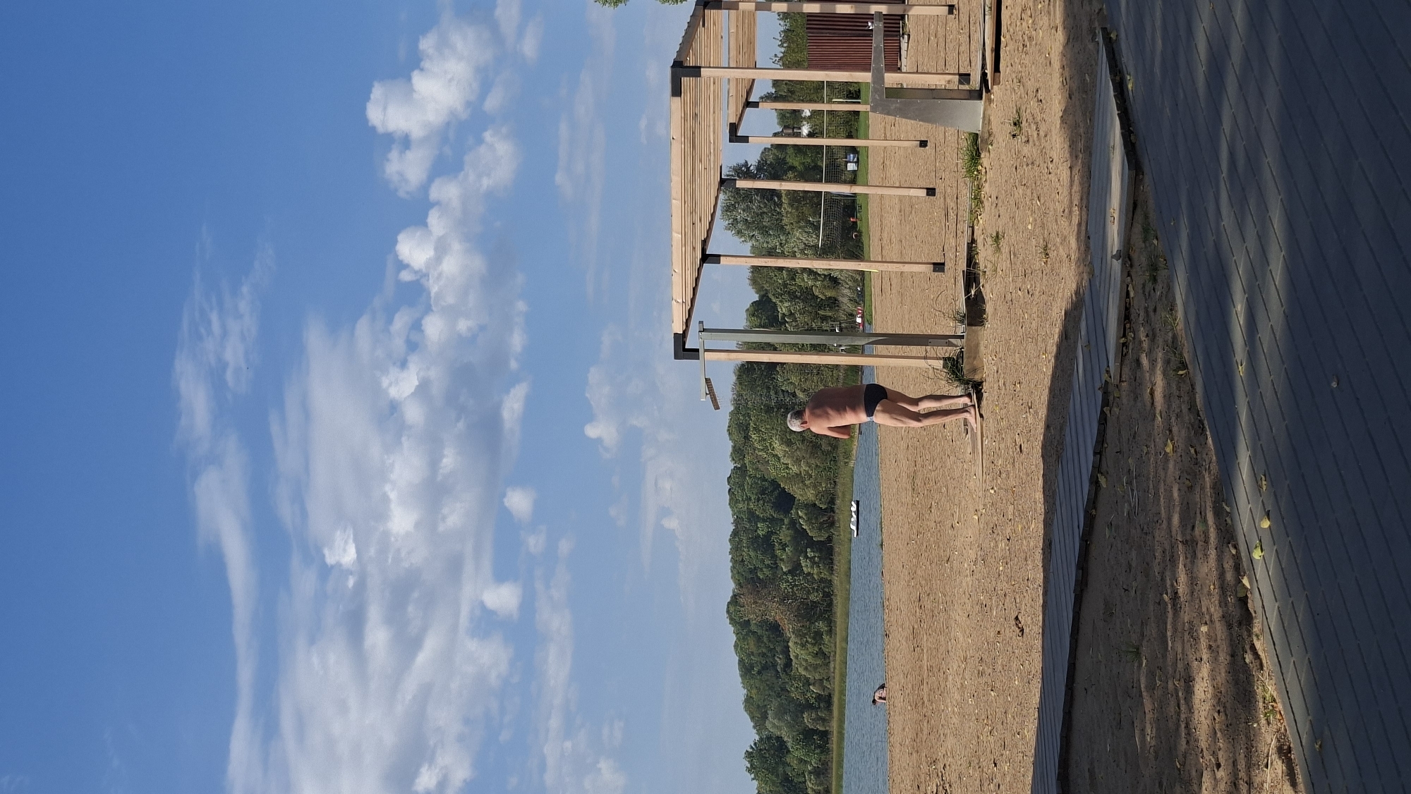 The image depicts a sandy beach area next to a lake, with a wooded area in the background. The sky is blue with scattered white clouds. In the foreground, a man in black swim trunks is standing under an outdoor shower. There is a wooden canopy structure behind him. Further back, there are people in the lake. Boardwalks are laid out on the sand leading towards the water. The scene appears to be a public beach area, possibly in the city of Myadzyel, Belarus, given the context provided.