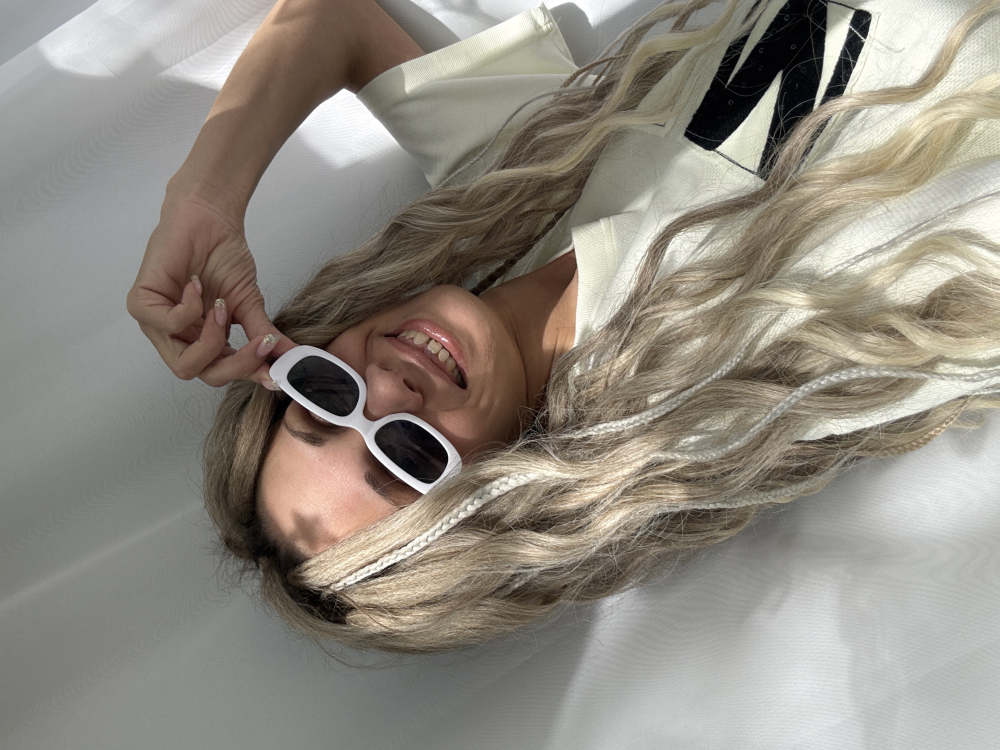 A woman lies on a white fabric surface indoors. She wears a white t-shirt with a large black letter "M" on it and white-framed sunglasses with black lenses. She is smiling and holding the sunglasses with her right hand. Her long, blonde hair with white braids is spread around her. The location is not discernible from the image.