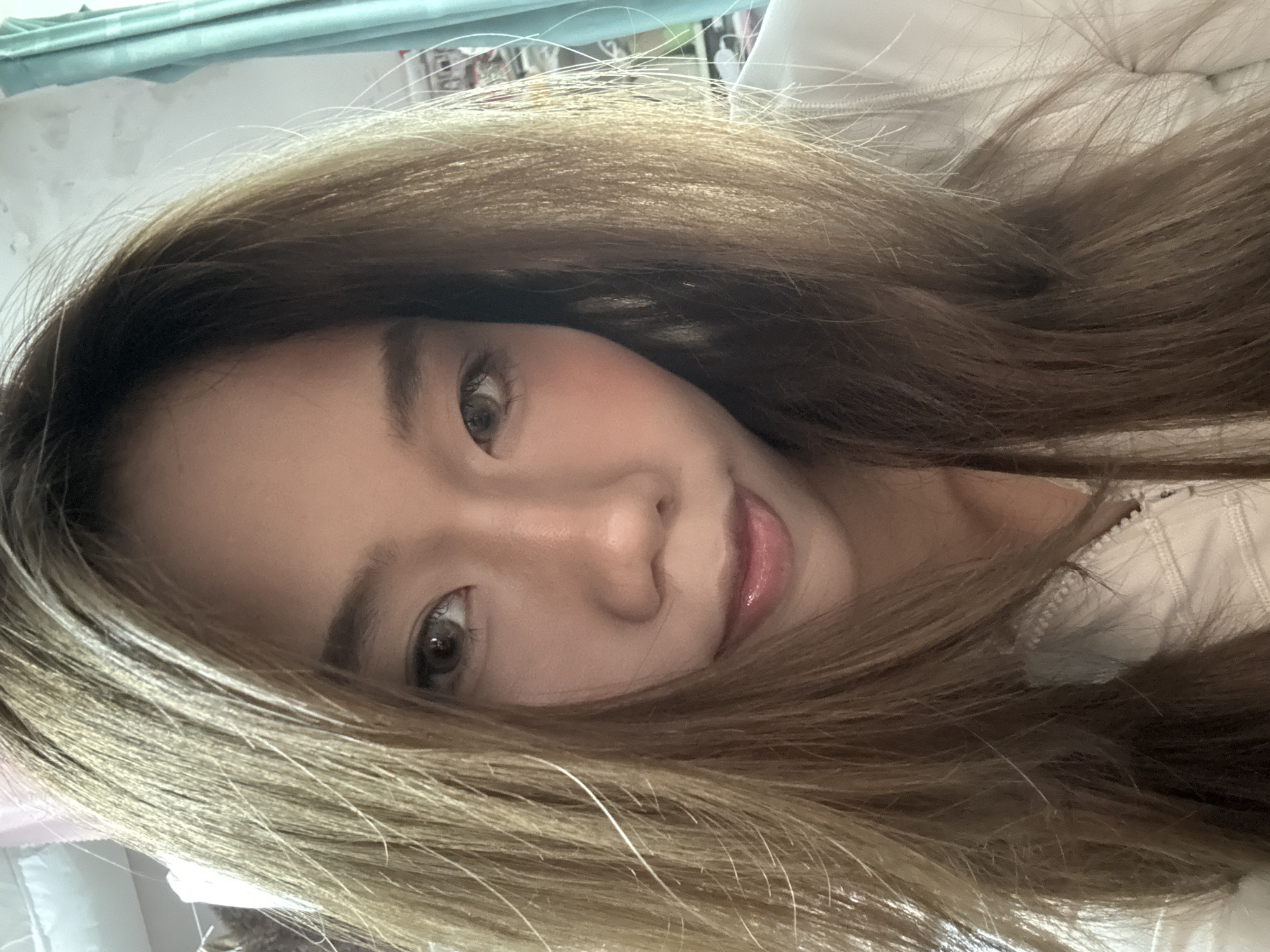 A close-up, angled selfie of a young Asian woman with long, light brown hair framing her face. Her face is fair with light makeup including pink blush and lip gloss. She has dark brown eyes. The background is blurred and appears to be indoors, with hints of a light-colored fabric or surface and a possible window. There is no indication of the location being Nan'an District, China.