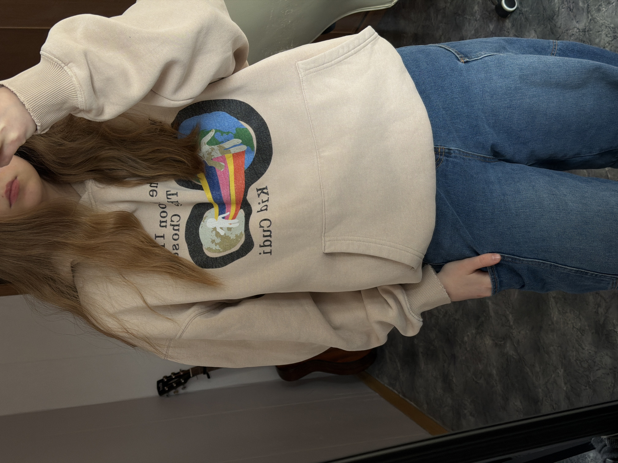 A person wearing a tan hoodie with a world graphic and blue jeans is photographed from an overhead angle. The jeans are a medium wash with visible seams and pockets. The hoodie has a front pocket and ribbed cuffs. The person's long brown hair is visible in the photo. The background includes a grey floor, a chair, and part of a guitar. The location is not identifiable as Perm, Russia.