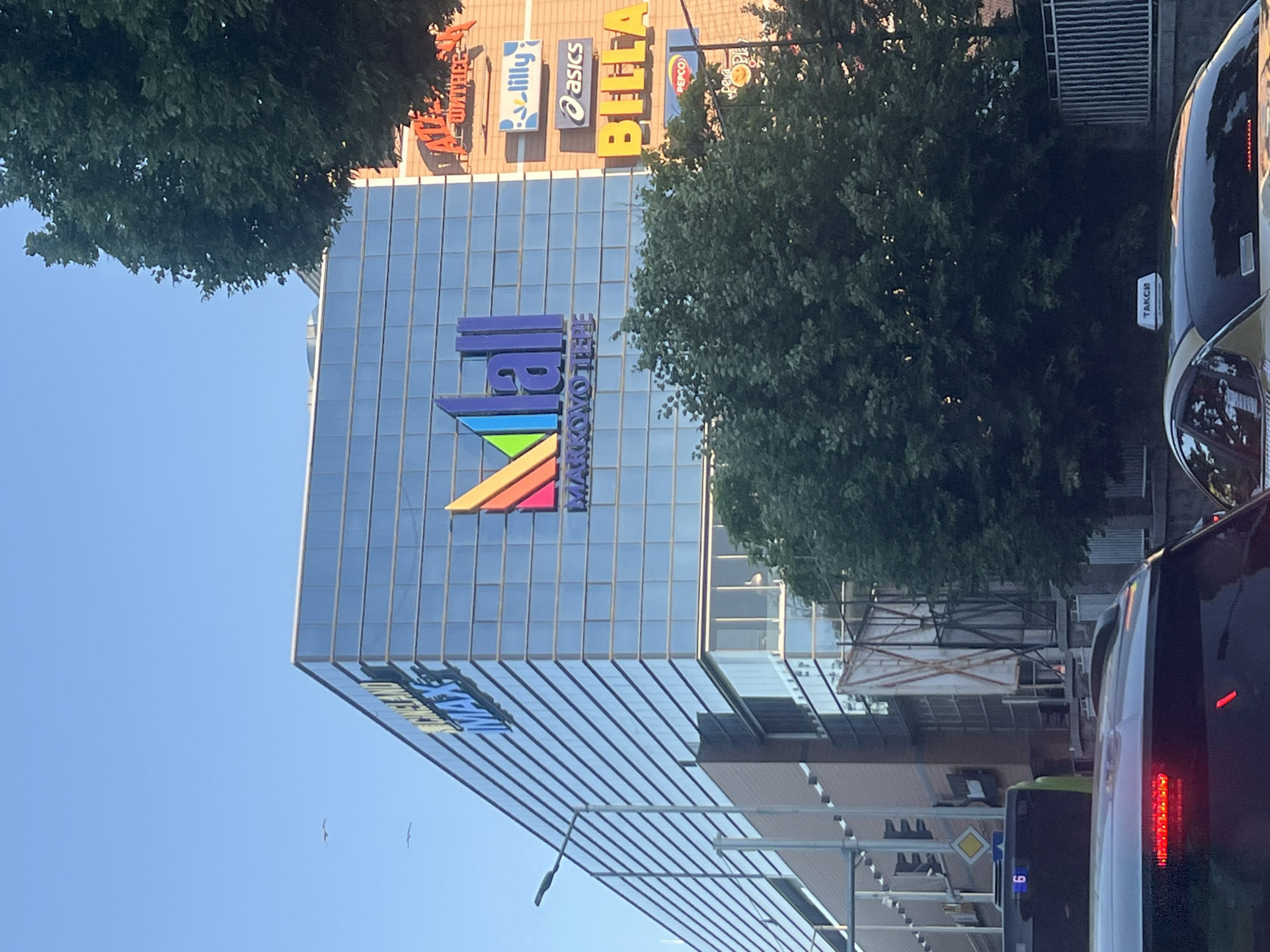 The image shows a building with a glass facade in Plovdiv, Bulgaria. The building has signage indicating "MARKOVO TEPE MALL" and other brands like Lilly, Asics, Bella, and Pepco. Street lighting is visible in front of the building. Traffic with cars and buses can be seen. A taxi with signage on the top is present. Trees are to the side of the building. The sky is clear and blue.
