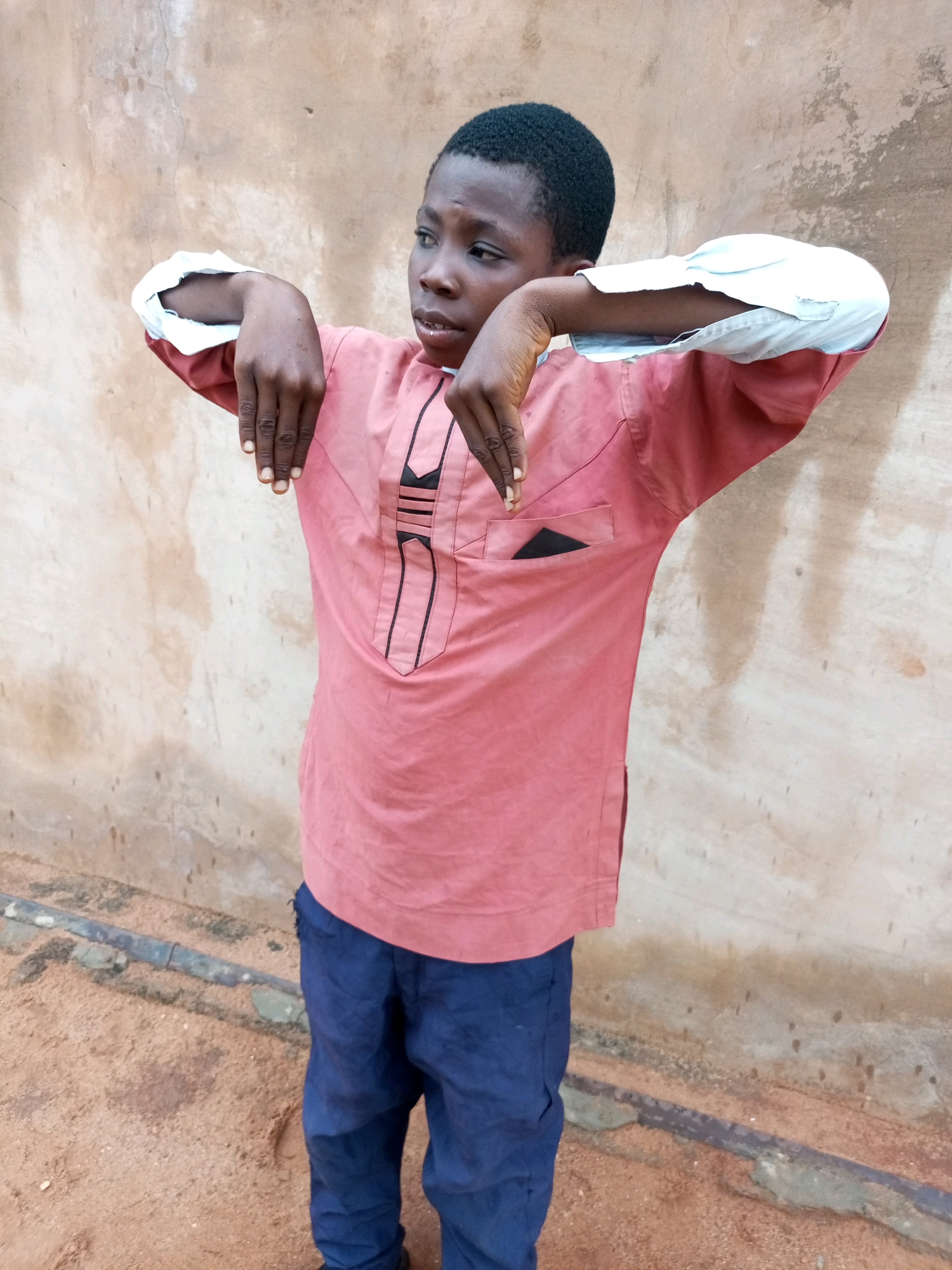 A young boy stands in front of a textured, light brown wall, his body angled to the left of the frame. He wears a coral-colored tunic with white cuffs and dark blue trousers. The tunic has black trim forming a decorative pattern on the chest and a small black pocket on the right side. His arms are bent, hands held up with fingers splayed, as if in a gesture of surprise or explanation. The ground beneath him is a reddish-brown, sandy earth, with a horizontal pipe or bar visible just above the ground. The wall behind him is uneven and weathered. The lighting suggests it is daytime, with soft, diffused light. The scene appears to be outdoors, possibly in a less developed area. The backdrop of the wall and ground suggests a utilitarian or rustic setting.
