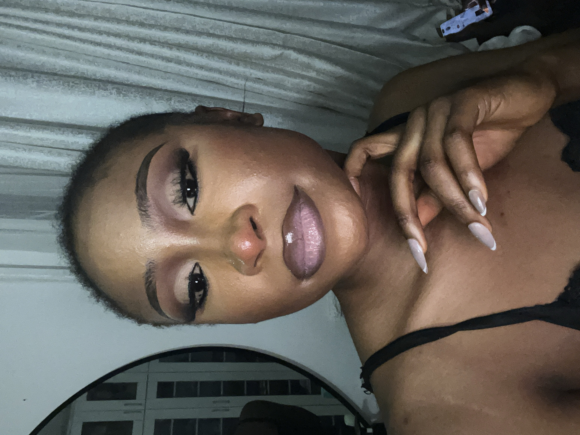 A woman with dark skin and full makeup is captured in a close-up, head-and-shoulders shot. She has on a black tank top with thin straps. Her right hand is positioned near her chin, with her fingers delicately touching her cheek. She has long, almond-shaped nails with a French manicure. Her lips are glossy and appear to be a mauve shade, while her eye makeup features a blend of browns and pinks, with dramatic eyeliner and false eyelashes. The background is a softly lit, light-colored curtain. A round mirror is visible on the left, reflecting a closet with mirrored doors. The scene is indoors, likely in a residential setting in Abuja, Nigeria.