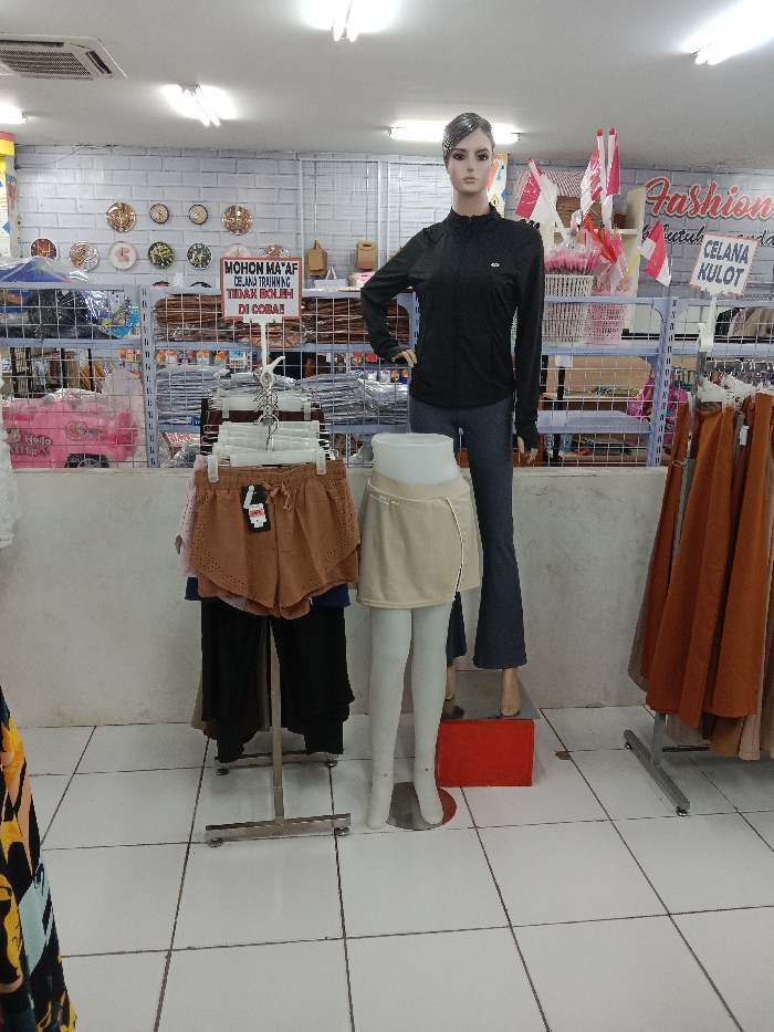 The scene depicts an interior view of a retail establishment, likely a clothing store, located in Langenharjo, Indonesia. The environment is brightly lit by overhead fluorescent fixtures on a white ceiling and features a white tiled floor and a white brick-patterned wall in the background.
In the foreground and midground, multiple clothing displays are arranged. Centrally, a full-body female mannequin with realistic features, short dark hair, and a neutral expression stands on a red rectangular block. It is dressed in a long-sleeved black athletic jacket with a zipper and dark blue flared trousers. To its left, a waist-down female mannequin with white legs displays a light beige athletic skirt with an asymmetrical wrap-style front, layered over what appear to be integrated leggings.
Adjacent to these mannequins, a silver-colored metal standing rack presents multiple pairs of brown perforated shorts with drawstrings, suspended on plastic hangers. Other dark-colored garments are visible beneath the shorts on this rack. To the far right, another silver-colored metal rack holds numerous garments, predominantly in shades of brown and orange, likely skirts or wide-leg pants.
In the background, behind the primary displays, several metal shelving units are visible. The leftmost unit contains pink "Hello Kitty"-branded items. Middle shelves are stacked with folded textiles or garments. The rightmost shelving unit displays small Indonesian flags (red and white) on sticks, alongside other packaged goods. A sign on this unit includes the word "Fashion" and "CELANA KULOT" (Culottes). Mounted on the background wall, to the left of the center, are several circular decorative plates or clocks. A white rectangular sign with red text, attached to a pole in the mid-left, reads "MOHON MA'AF CELANA TRAINING TIDAK BOLEH DI COBA," which translates to "SORRY TRAINING PANTS CANNOT BE TRIED ON." An air conditioning vent is visible on the ceiling in the upper left quadrant of the image. No individuals are present, and no actions or interactions are occurring; the scene is a static retail presentation.
