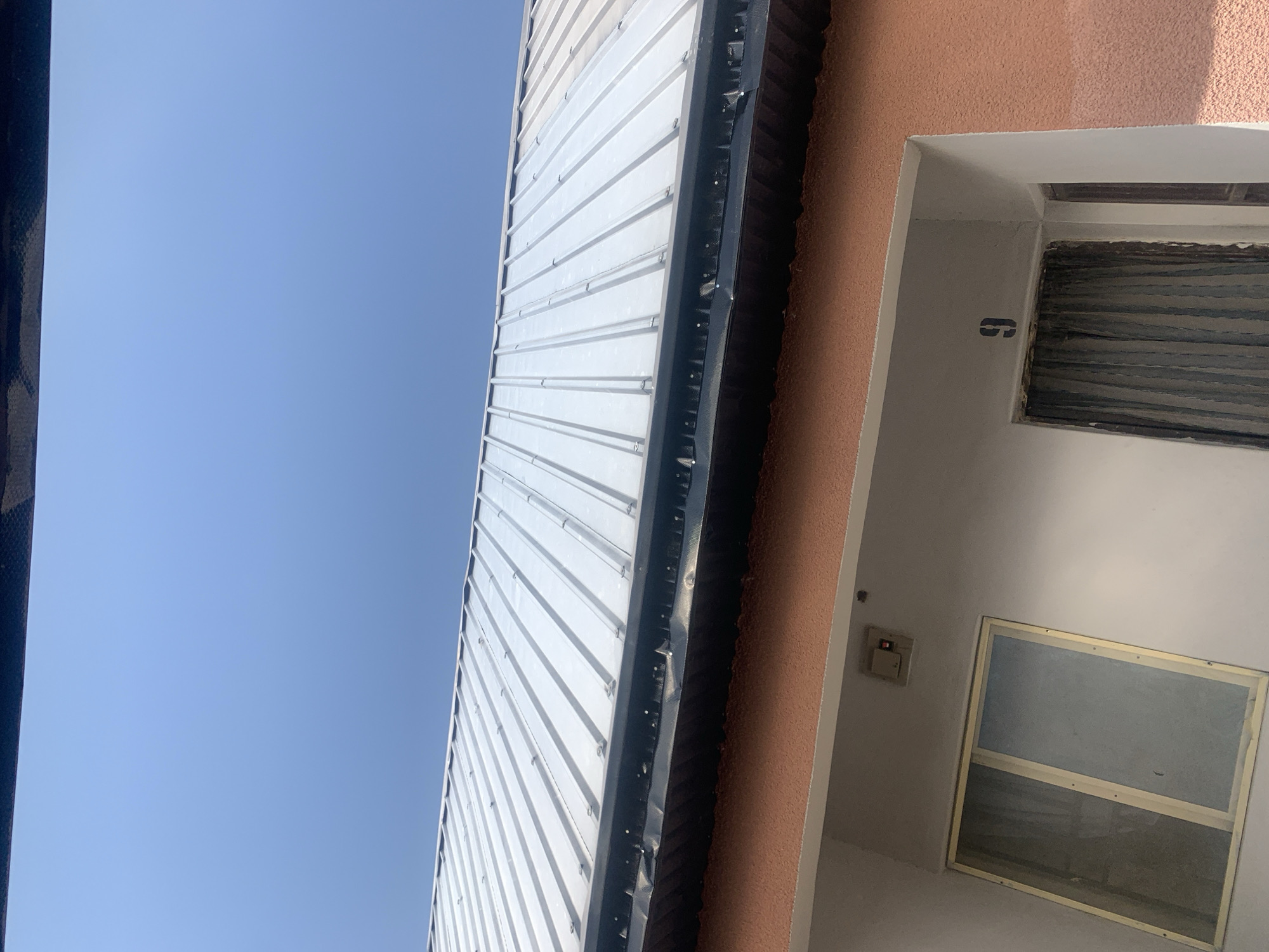The image captures a close-up, low-angle view of a building's facade and roof under a clear blue sky. The roof is constructed of corrugated metal, likely tin, with visible fasteners. A dark-colored material, possibly a gutter liner or flashing, runs along the edge of the roof. Below the roofline, the building features an orange-textured wall. On this wall, there is a white framed window on the left, appearing to have a gray tint. To the right of the window, a doorway is visible, with dark curtains drawn across it. A black stencil number "9" is displayed above the doorway. A small electrical box with a red light and a switch is mounted on the wall above the window. This scene is located in Jauro Yinu, Nigeria.