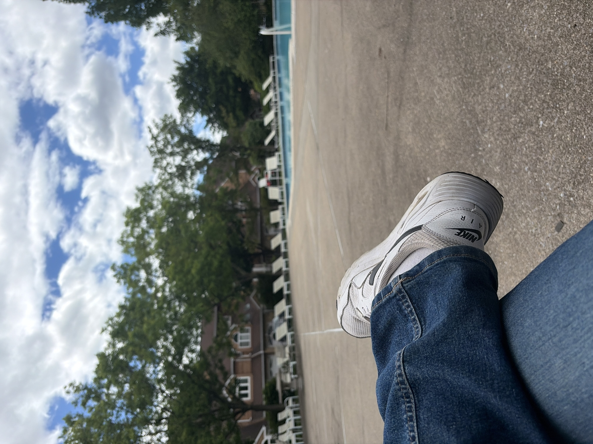 A person wearing blue jeans and white Nike Air sneakers is sitting on a concrete surface. In the background is a swimming pool with white lounge chairs, a building, and green trees under a partly cloudy sky. The location appears to be an outdoor recreational area, possibly a pool or club facility in Edison, New Jersey, United States of America.