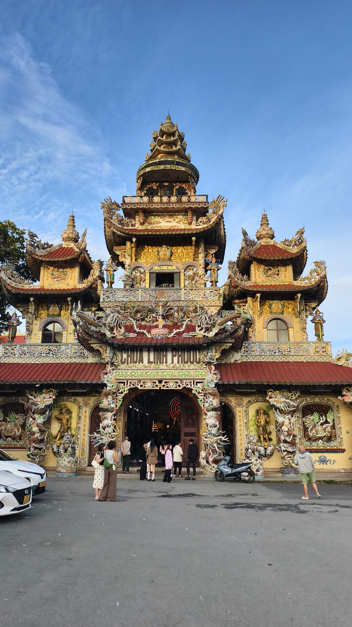 The image shows the facade of Chua Linh Phuoc, a Buddhist temple located in Xuân Thành, Vietnam. The temple is adorned with colorful mosaics and intricate dragon sculptures. The building features multiple tiers of pagoda-style roofs and ornate detailing. In front of the temple, several people are standing, some near parked vehicles, including a white car and a motorbike. The sky above is blue with scattered clouds.