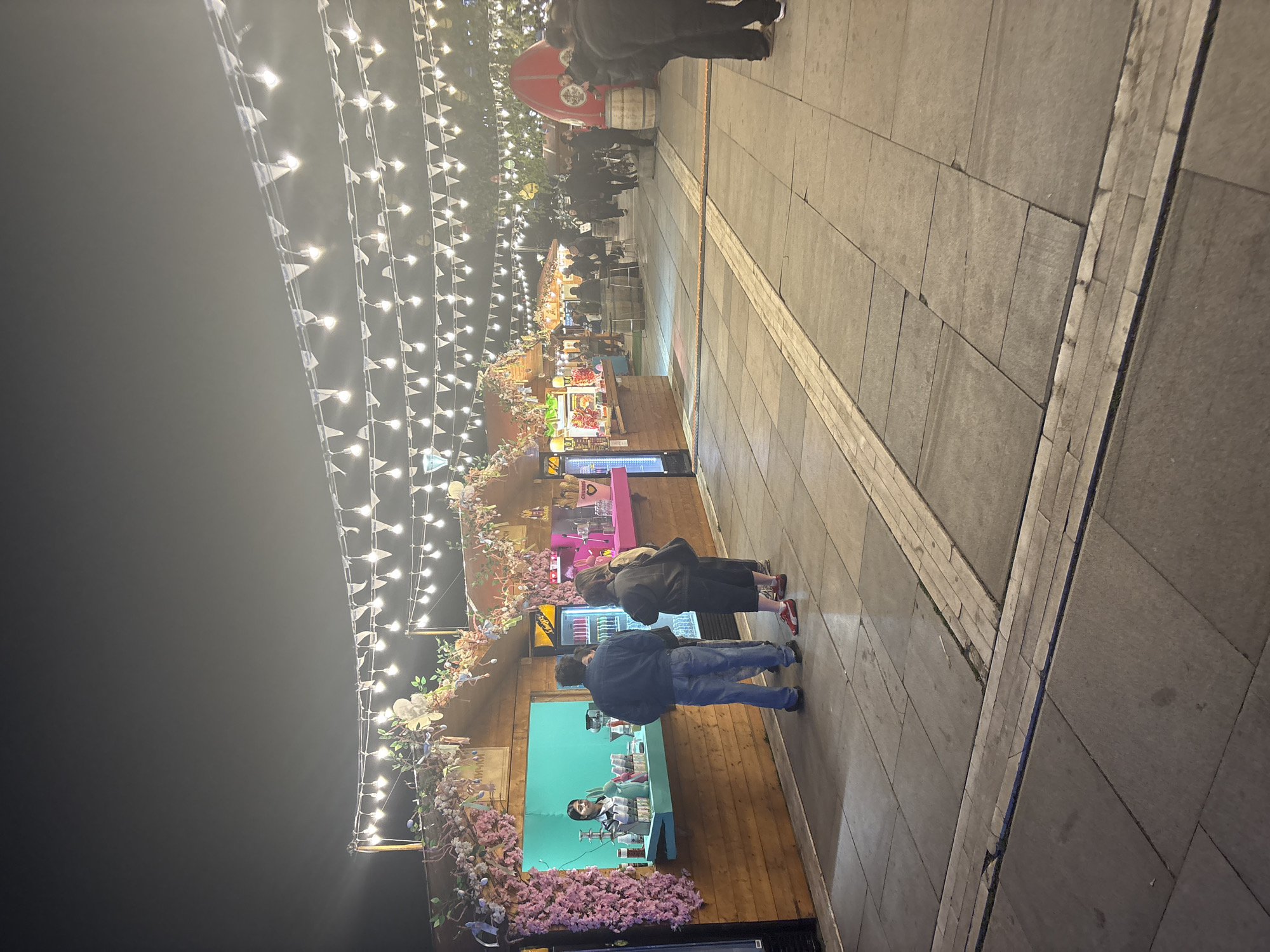 This image depicts a lively outdoor night market or fair in Sofia, Bulgaria. The scene is illuminated by numerous strings of small white flags and bright light bulbs, creating a festive ambiance against the dark night sky.
Several people are visible along a paved walkway. In the foreground, two individuals stand near a market stall, facing its counter. One wears a dark jacket and blue jeans, while the other is in a dark coat. A vendor is partially visible behind the counter of the nearest stall, which is adorned with pink cherry blossom decorations. Further along the walkway, more people are scattered, some gathered near other stalls, suggesting browsing or purchasing activities.
The market stalls are constructed from wood and are brightly lit, displaying various items. A distinctive red, mushroom-shaped structure is visible in the mid-ground, with wooden barrels placed beside it. The ground is paved with large, grey square tiles, featuring lighter-colored decorative strips. A sign on a distant stall displays text that appears to be in Cyrillic, possibly reading "АМ ЧИЙКН" (AM CHICKEN). The overall atmosphere is bustling and social, indicative of an evening event or gathering.