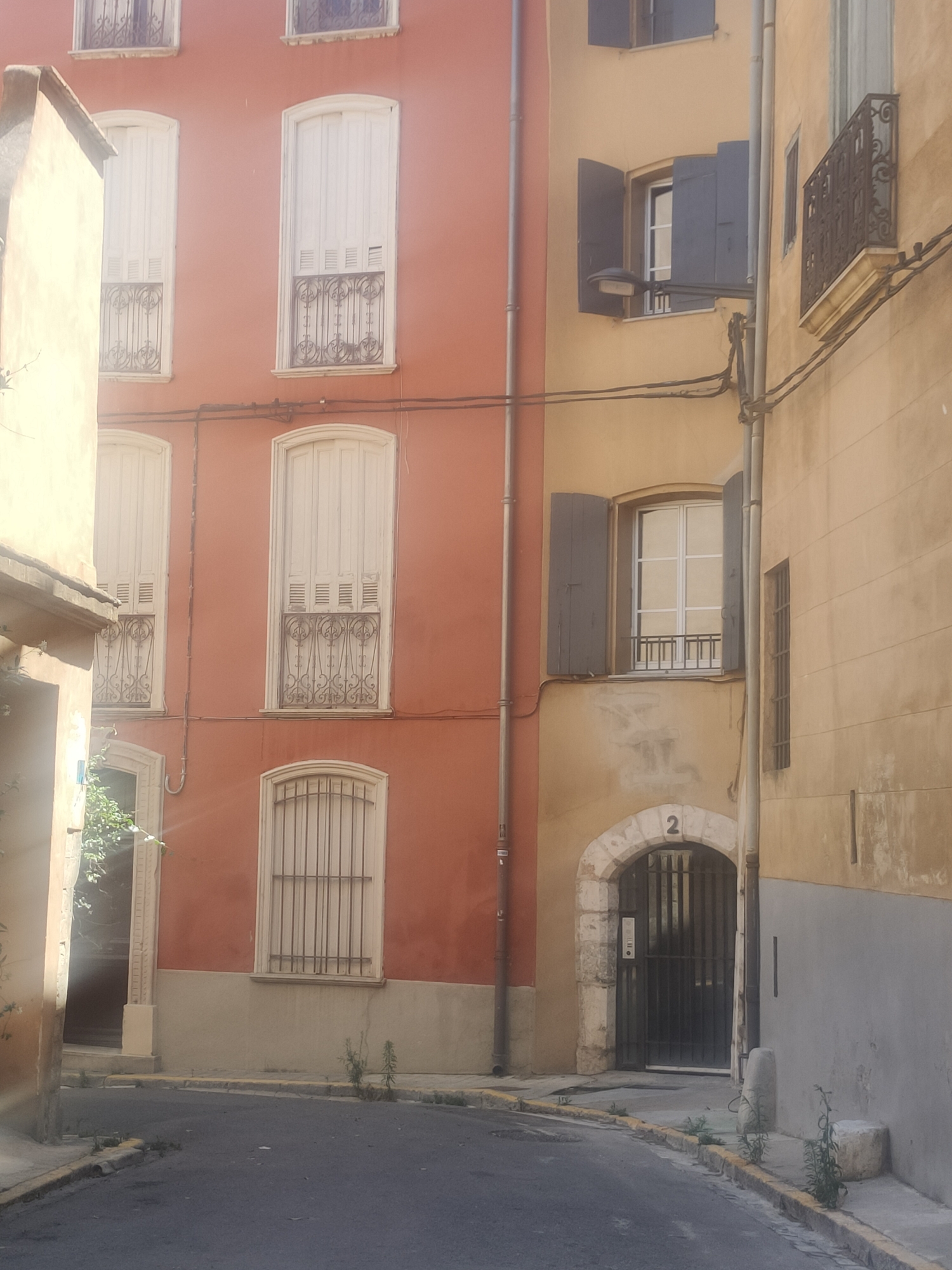 This is a photograph of a narrow street in Perpignan, France, lined with multi-story buildings. The buildings exhibit a mix of architectural styles and colors, with one prominent facade painted in a warm, terracotta-orange hue and an adjacent one in a muted yellow. The buildings feature numerous windows, some with white shutters and ornate ironwork, others with dark shutters partially open, revealing glimpses of interior spaces. A few windows are barred.
The street itself is paved with asphalt and bordered by a curb with yellow markings. Sparse vegetation, including small weeds and shrubs, grows along the roadside and in the cracks of the pavement. A few concrete elements, possibly bollards or low walls, are visible on the sidewalk.
The setting appears to be a residential or mixed-use area within the city. The lighting suggests it is daytime, possibly late morning or early afternoon, with sunlight casting some shadows. The scene is quiet and appears uninhabited, with no people or vehicles visible. The overall impression is one of historic European urban architecture, with a slightly weathered but charming feel. A house number "2" is visible above an arched doorway.