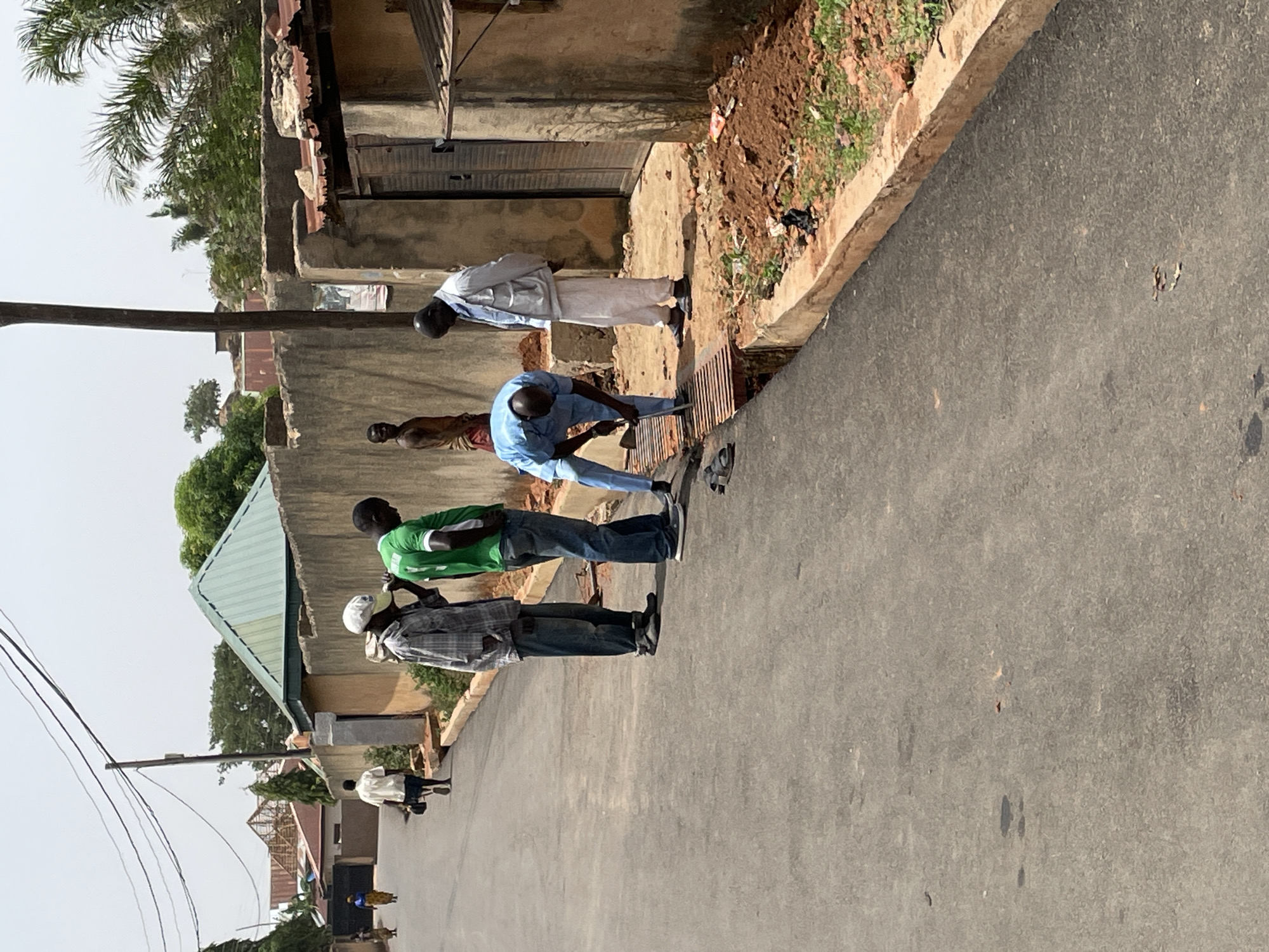 The image shows a street scene in Jos, Nigeria. The street is paved and lined with buildings of varying construction. At least eight people are visible, including a group of men standing near the edge of the road, some of whom appear to be working or interacting with a drain. Other people are walking further down the road. The buildings appear to be residential or commercial, with some showing signs of age or incomplete construction. The environment suggests a relatively populated urban area in Jos. Vegetation, including trees and shrubs, is present in the background and alongside the buildings. Power lines are visible in the sky.