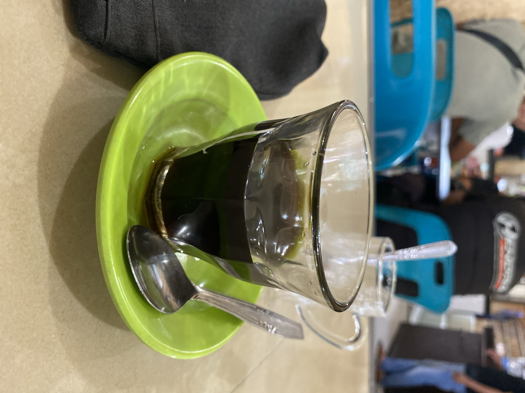 A close-up shot captures a glass of dark coffee resting on a bright green saucer. A small silver spoon lies on the saucer, partially extending from the coffee cup. In the background, slightly out of focus, are the blurred figures of people and blue chairs, suggesting an indoor seating area, possibly a cafe or restaurant. The overall impression is a still life of a beverage, set in a public space. The scene is consistent with a cafe in Lhokseumawe, Indonesia.
