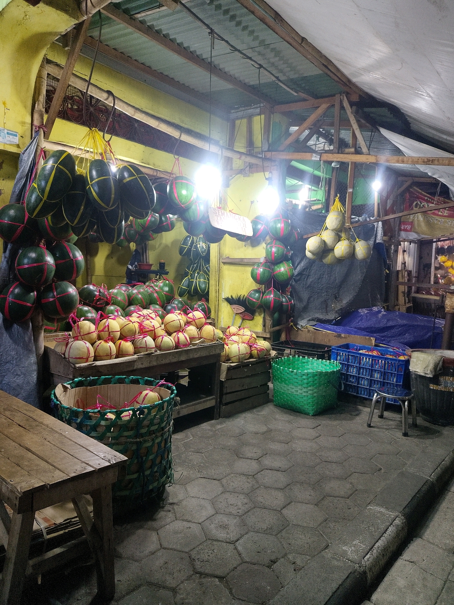 This is a photograph of a fruit stand in Mojokerto, Indonesia, possibly at night given the artificial lighting and the dark outdoor environment. The stand appears to be set up outdoors under a makeshift roof structure constructed from corrugated metal and wood beams, draped with a white tarp on one side. The ground is paved with hexagonal concrete tiles.
The primary subjects are a large quantity of fruits, predominantly watermelons and what appear to be cantaloupes or similar melons. The watermelons, many of which are dark green with yellow bands, are displayed in several ways: hanging from hooks on the ceiling of the stand, piled on wooden tables, and in large woven baskets. Some of the hanging watermelons are wrapped in netting or string. The rounder, lighter-colored melons are also displayed on wooden tables, some in crates and baskets, often with decorative pink string tied around them.
In the background, there are more fruits, along with various crates, tarps, and possibly other market stalls or structures. A small blue plastic stool is visible near a blue plastic crate filled with more items. There are no people clearly visible in the image, suggesting the vendor may be absent or not in the frame. The overall impression is of a busy, yet organized, fruit market stall, likely operating in the evening.