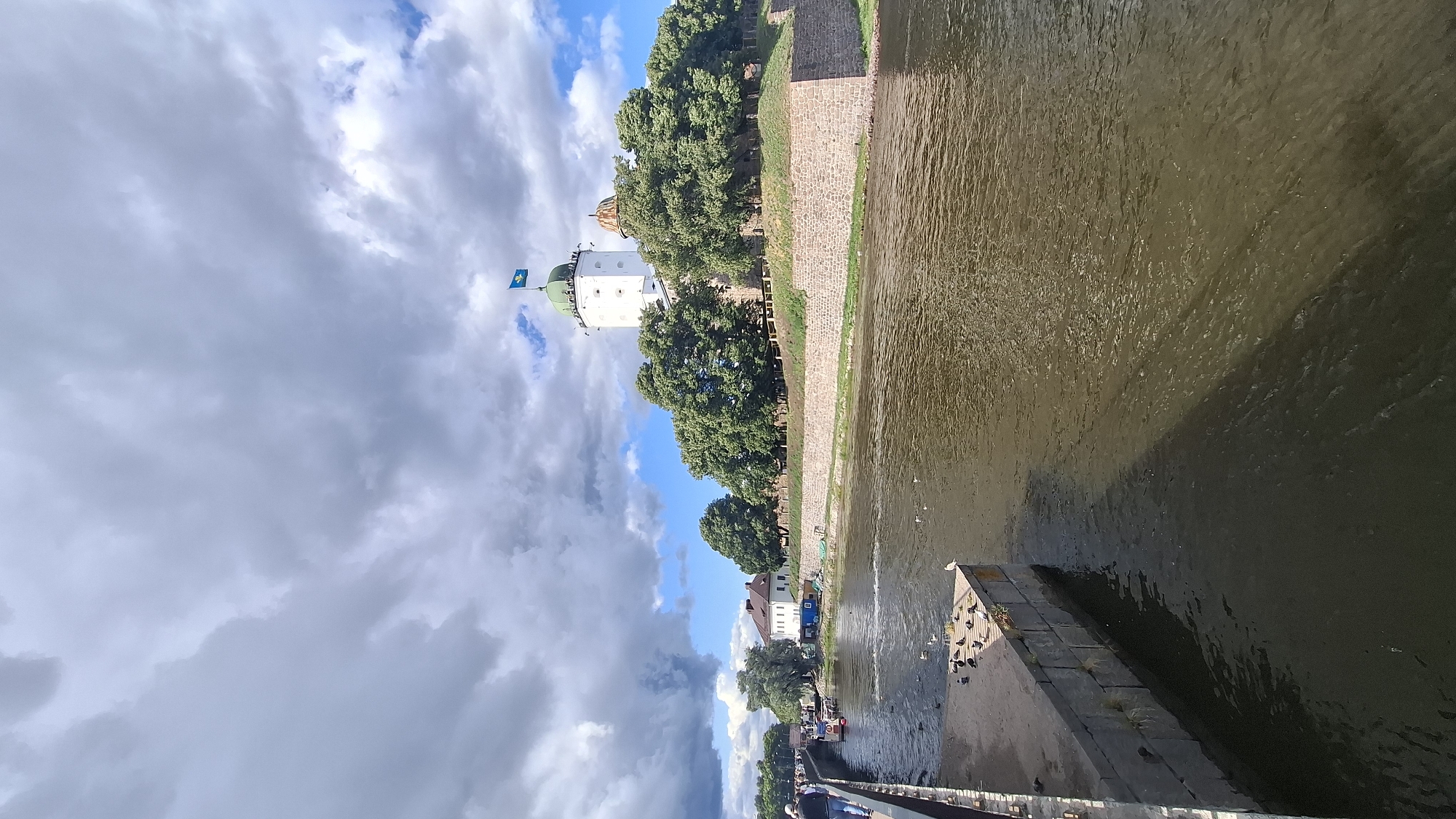 A wide-angle, high-angle shot captures the Vyborg Castle complex in Vyborg, Russia, on a partly cloudy day. The imposing castle, with its white main tower topped by a green dome and a blue flag, stands on a slight rise overlooking a wide, calm river. The castle walls, made of rough-hewn stone, form a protective barrier around the green, tree-covered grounds.
In the foreground, a concrete embankment lines the river on the left side. Several pigeons are gathered on this embankment, some pecking at the ground, others resting. The river itself is a murky brown color, with ripples disturbing its surface. A long shadow, cast by the embankment, stretches across the water.
Further along the river, on the left bank, a pedestrian walkway is visible, with a few people strolling. A small boat is docked near the walkway. In the background, several buildings, including a white structure with a red roof, are situated behind the trees. The sky is a mix of dramatic gray clouds and patches of bright blue. The overall scene evokes a sense of historical grandeur and natural beauty.
