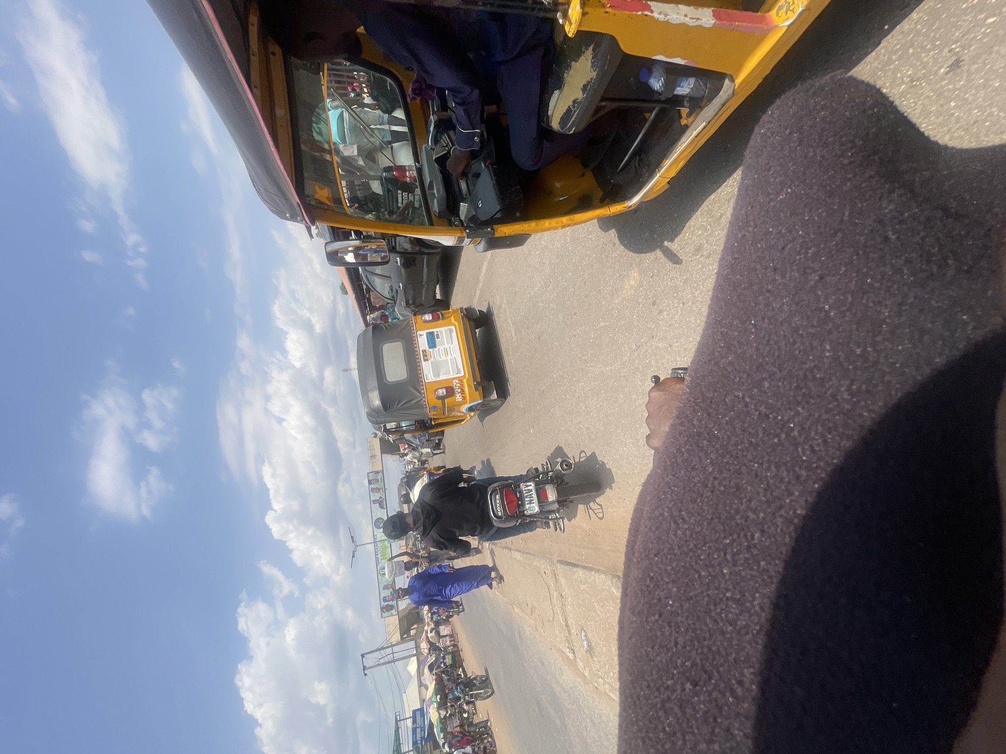 A slightly high-angle, wide shot in Lokoja, Nigeria. A street displays a mixed array of vehicles: a yellow and black auto rickshaw ("keke"), motorcycles, and black passenger cars. The road is lined with shops with colorful signage. Sky is partly cloudy. A person's leg wearing dark clothing is in the right foreground.