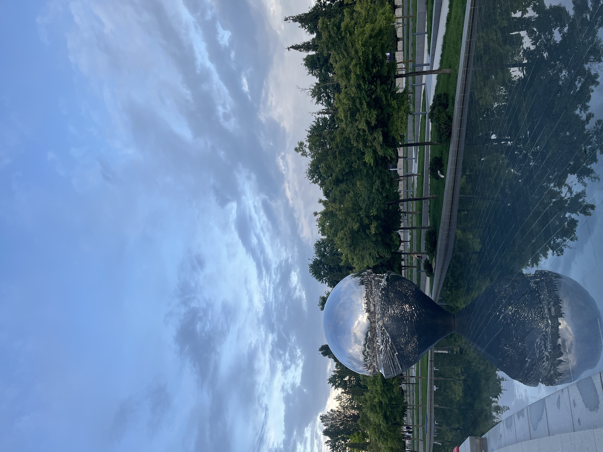 The image shows a landscape in Krasnodar, Russia. In the foreground, there is a reflective sculpture that captures the surrounding environment in its surface. The sky is filled with a mix of blue and gray clouds. In the background, a row of trees separates the sky from a water feature where the trees, sky, and sculpture are reflected.