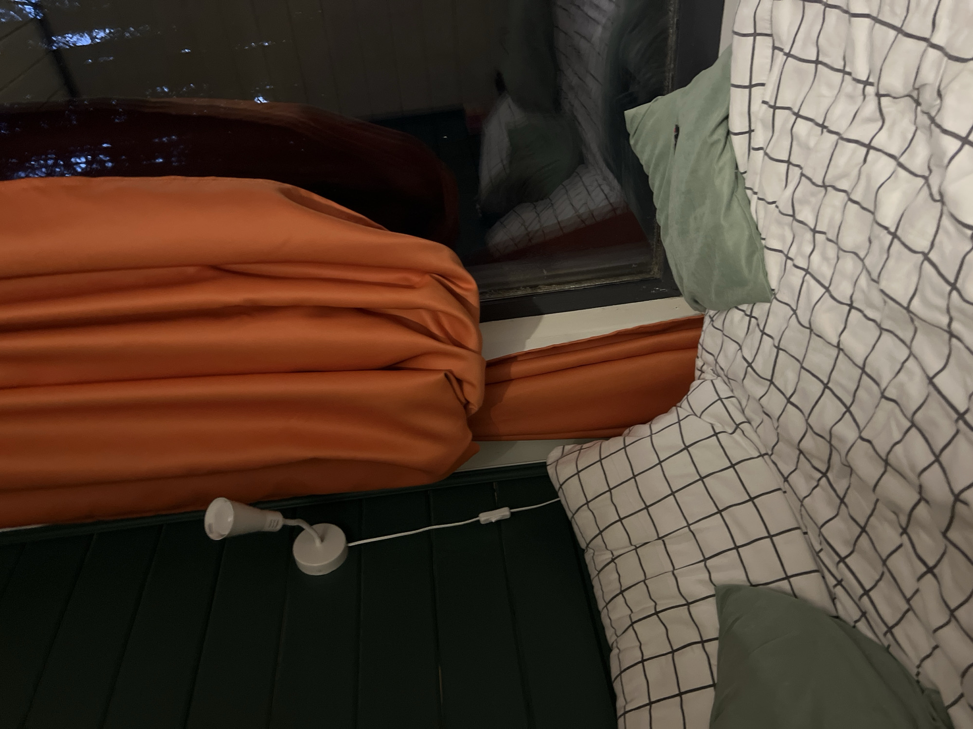 The image shows an indoor scene, possibly a bedroom. An orange drape or curtain is prominently displayed on the left. A bed with a white and black checkered pattern bedspread and two green pillows is on the right. A small white lamp is attached to the green wall. The location in the city of Polazna, Russia, cannot be determined from the image.