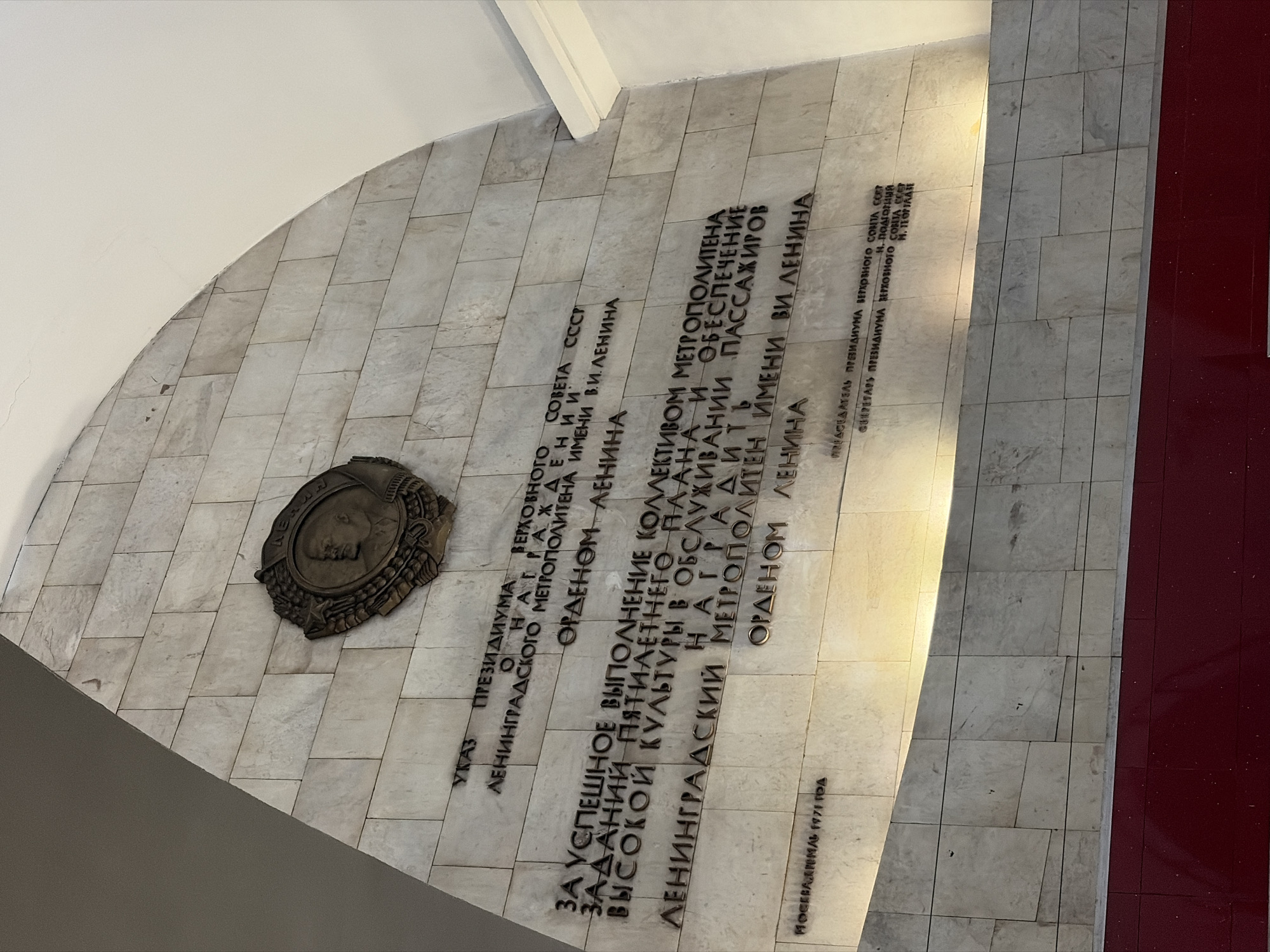 The image shows a stone plaque mounted on a wall, likely located within a metro station in Saint Petersburg, Russia. The plaque features an oval-shaped bronze medallion with a profile portrait, possibly of Vladimir Lenin. Below the medallion, an inscription in Russian is engraved in block letters, citing a decree to award the Lenin Order to the Leningrad (Saint Petersburg) Metro, named after Lenin. The inscription states the reason for the award is for successful fulfillment of the five-year plan and high standards of passenger service. The date of the decree appears at the bottom of the inscription, "Moscow, Kremlin, 1971." The plaque is affixed to a wall tiled with light-colored, rectangular stone or ceramic tiles. The lighting is uneven, with the right side of the image brighter than the left. A section of dark red flooring is visible along the right edge of the image.