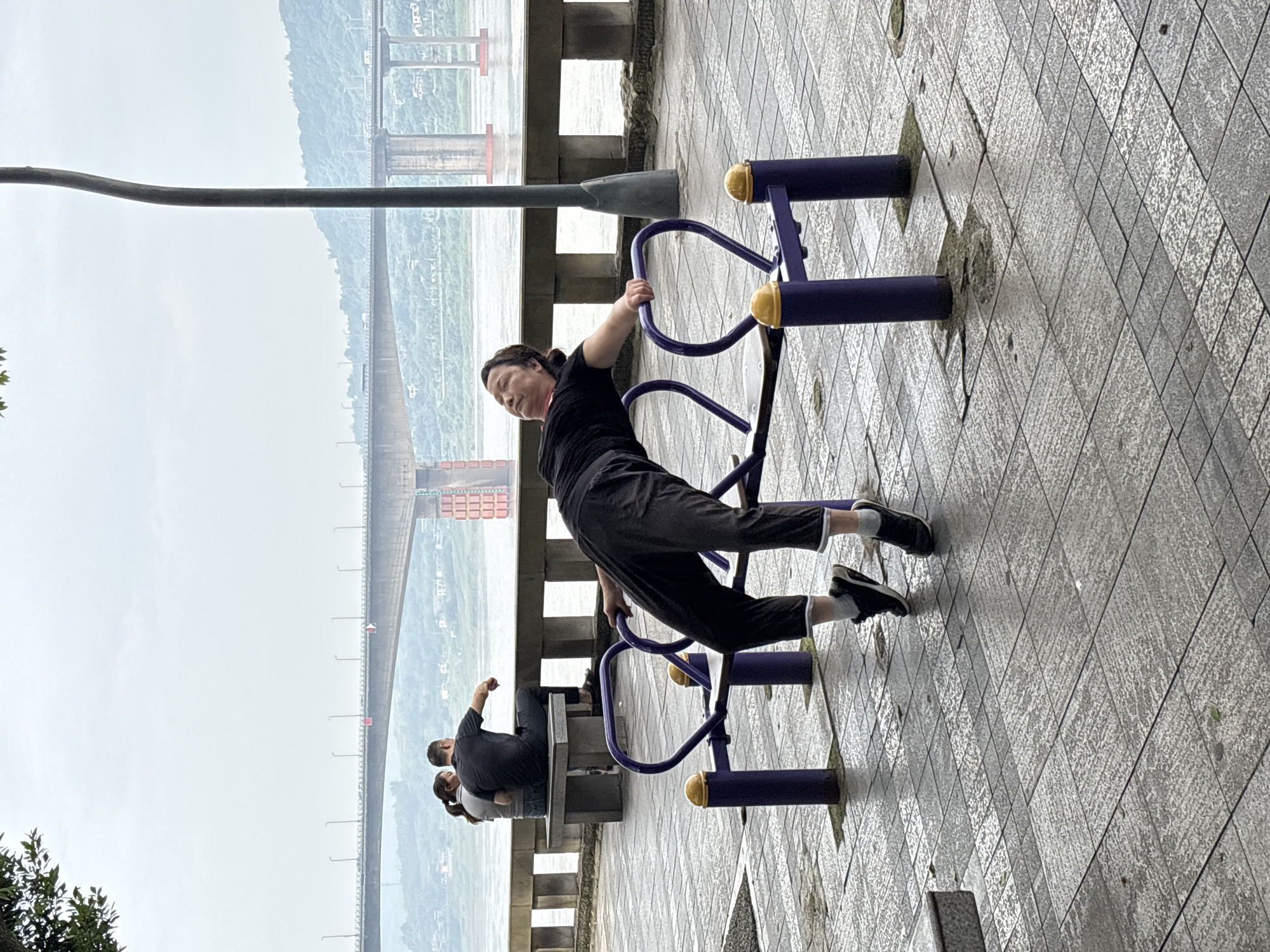 A woman is seen relaxing on a purple and yellow exercise bench by a river, with her body arched backward and her head tilted up. She is wearing a black t-shirt and black pants. The exercise equipment is on a tiled walkway. In the background, a bridge spans the river, and on the opposite bank, there are hills and some buildings. Further back, another couple is seated on a stone bench. The sky is overcast. The scene appears to be in the city of Jiangjin, China, a district of Chongqing.