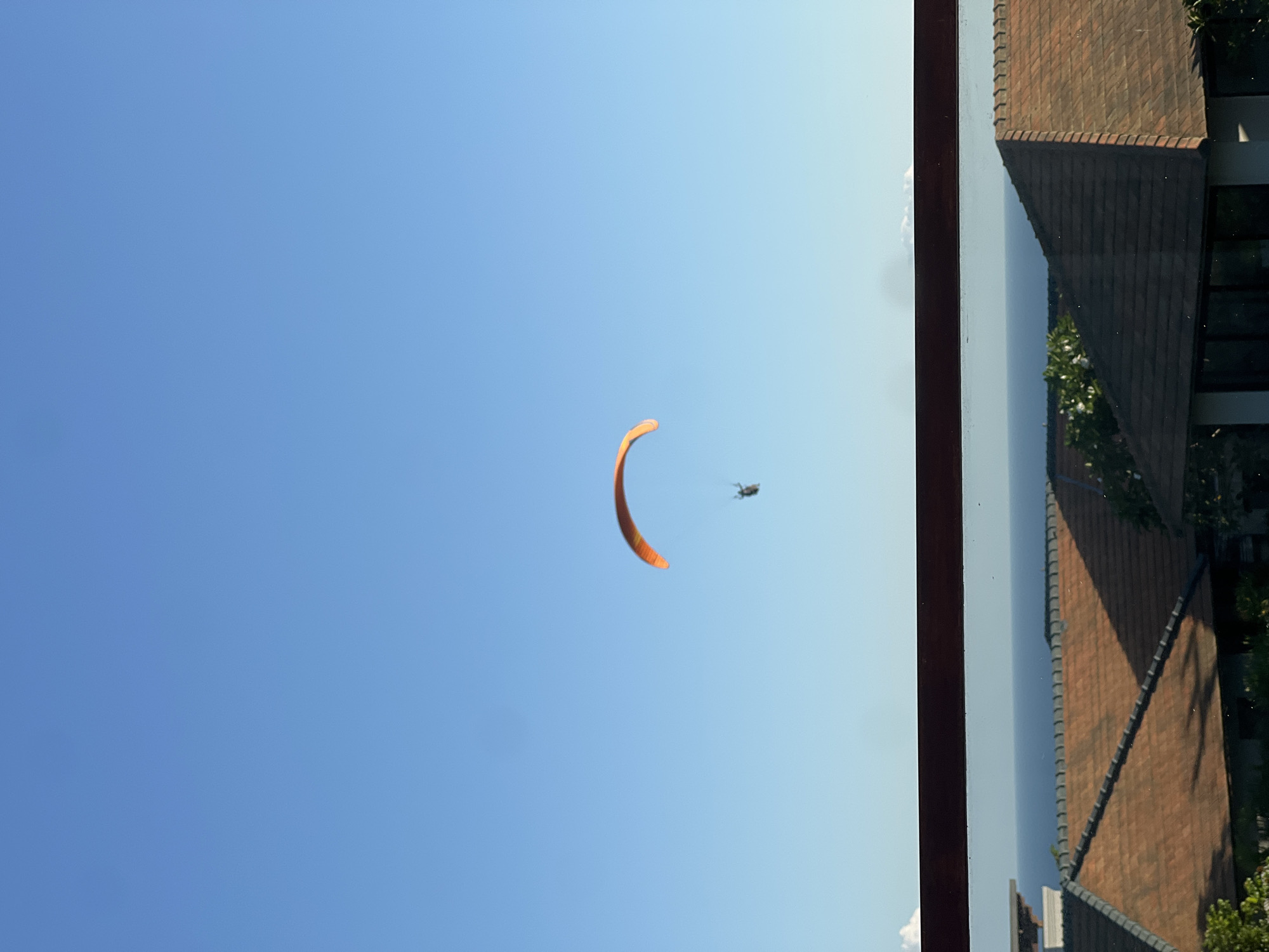 The image displays a paraglider soaring in a bright, clear blue sky, viewed from an indoor perspective looking outwards. A vivid, arc-shaped orange paraglider wing is prominently featured in the upper-middle section of the sky. A small, dark figure, presumably the paraglider pilot, is visible directly beneath the wing.
On the right side of the frame, a dark, thick vertical structure suggests a window or door frame, through which the scene is observed. Beyond this frame, the top portion of a building is visible, featuring a steeply pitched roof with reddish-brown tiles and a darker facade, possibly brick or stone. Some green foliage and dark shadows are also present near the building, hinting at surrounding greenery.
The activity taking place is paragliding. The clear, cloudless sky and bright illumination indicate that it is daytime, likely morning or afternoon, with excellent weather conditions for outdoor aerial sports. There is no visible text in the image. The setting appears to be an outdoor scene viewed from a building.