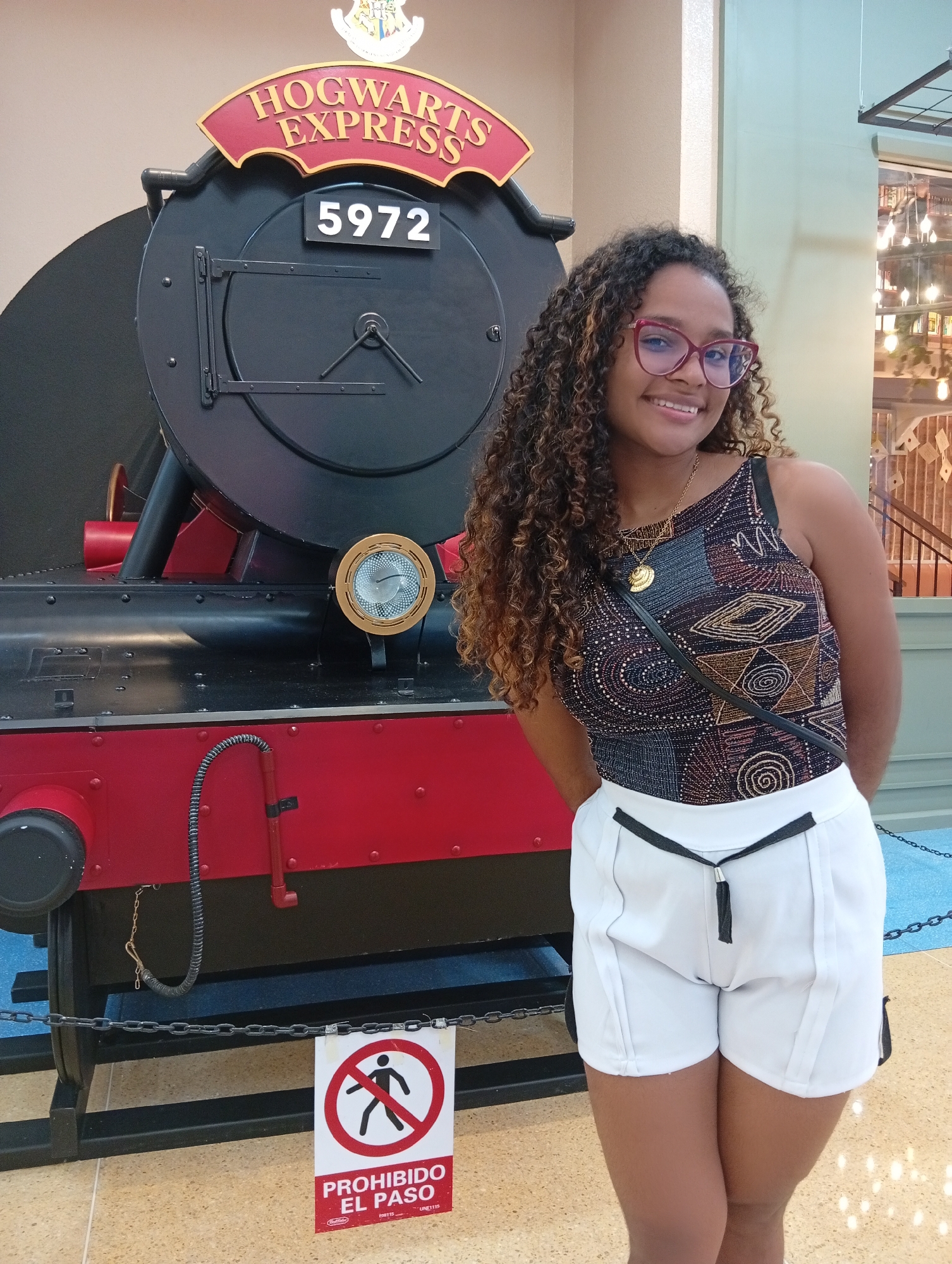 A young woman is standing next to a replica of the Hogwarts Express train. The woman has curly brown hair, is wearing red eyeglasses, a patterned top, and white shorts. The train is black and red with the words "Hogwarts Express" and the number "5972" displayed on it. A sign that says "PROHIBIDO EL PASO" is in front of the train. The scene appears to be indoors, possibly at a Harry Potter-themed exhibit. The location is presumed to be Porlamar, Venezuela.
