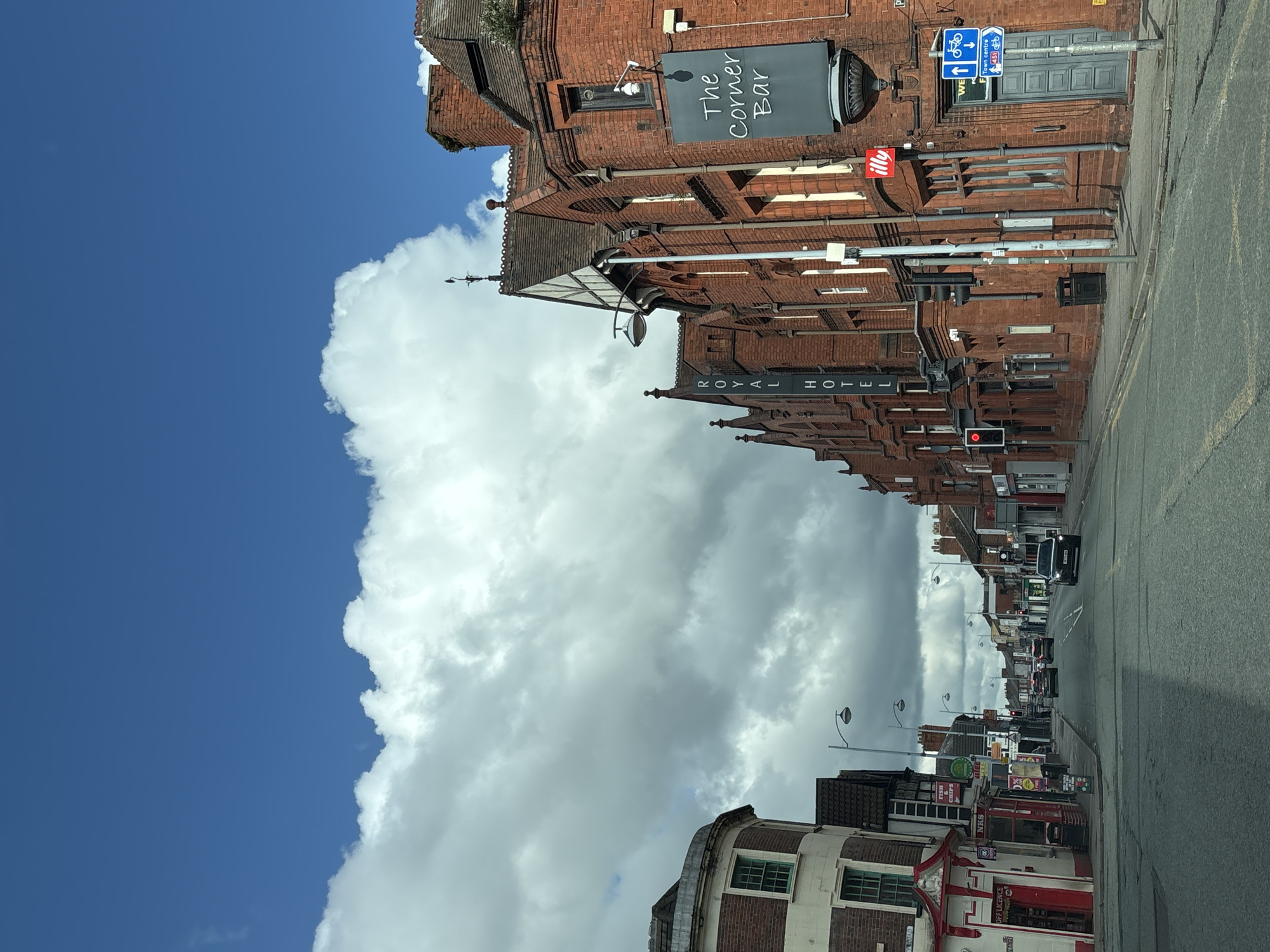 The image shows a street scene in Crewe, United Kingdom. The view is of a street lined with brick buildings. "The Corner Bar" and "Royal Hotel" are identifiable establishments. A large cloud formation dominates the sky next to clear blue. Streetlights line the road, and there are traffic signals visible. Several cars are on the street. Shopfronts with signage are also visible. A street sign with "Town Centre" and a bicycle symbol is present.
