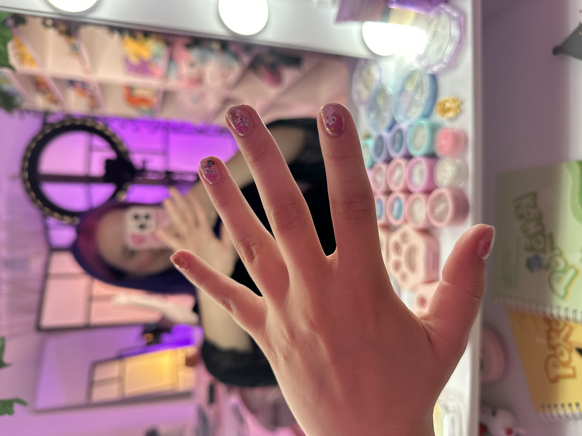 The image shows a person's hand with manicured nails, each decorated with small, cartoon-like figures. The background is a mirror reflection of a room with purple lighting, a ring light, and shelves filled with colorful items. In the foreground, below the hand, are rows of small containers, possibly for nail polish or makeup, and some notebooks. The scene appears to be a personal vanity area. There is no indication of the city of Oviedo, Spain.