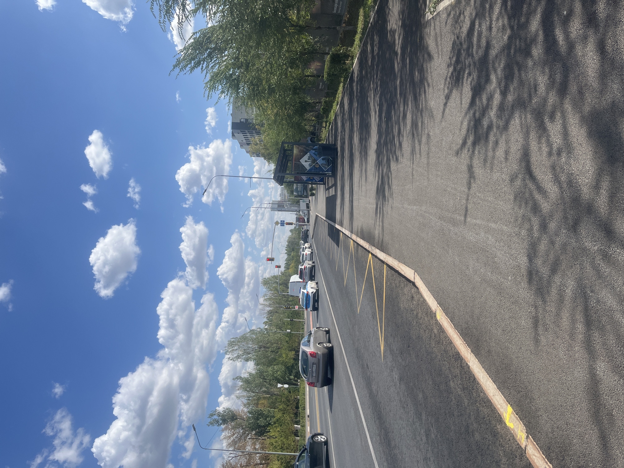 The image depicts a sunny day in Astana, Kazakhstan, along a multi-lane road. The sky is a bright blue with scattered white cumulus clouds. On the right side of the road, there is a sidewalk with trees casting shadows on the asphalt. A bus stop shelter with graphic advertisements and a trash bin is visible. Beyond the trees, modern apartment buildings rise into the sky. On the left side of the road, cars are driving in both directions. A silver sedan is in the foreground, followed by other vehicles, including a white van with police markings and several passenger cars. The road has yellow markings on the curb, indicating a no-parking zone. Traffic lights are visible in the distance. The overall scene is one of urban infrastructure under a clear sky.