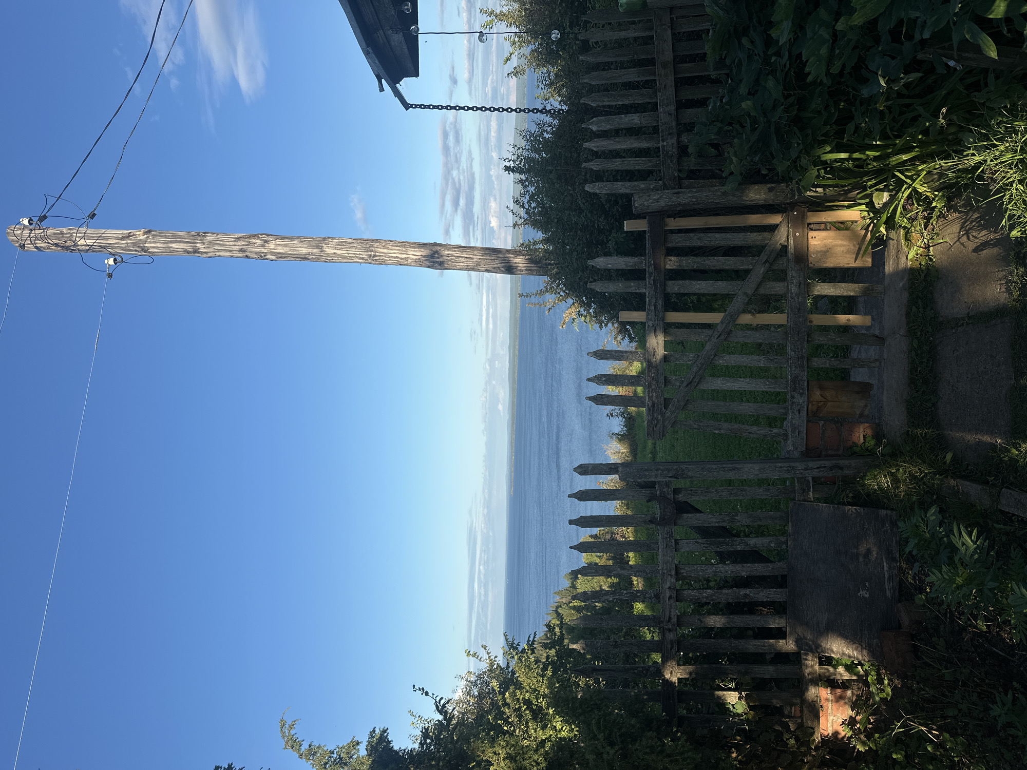 The image captures a serene outdoor scene, likely in the village of Galievo, Russia, on a clear day. In the foreground, a weathered wooden fence with pointed pickets encloses a garden area. A gate, partially ajar, reveals a glimpse of lush green grass beyond. To the left, a dense cluster of trees and foliage casts shadows on the ground, while to the right, a rough-hewn wooden utility pole stands tall, supporting electrical wires against the bright blue sky dotted with a few wispy clouds. Beyond the fence and trees, a vast expanse of calm blue water stretches towards a distant horizon, where a faint line of land is visible. A rustic wooden structure, possibly a shed or part of a house, is visible on the far right, with a chain hanging from its eaves. The overall atmosphere is one of peaceful, rural tranquility.