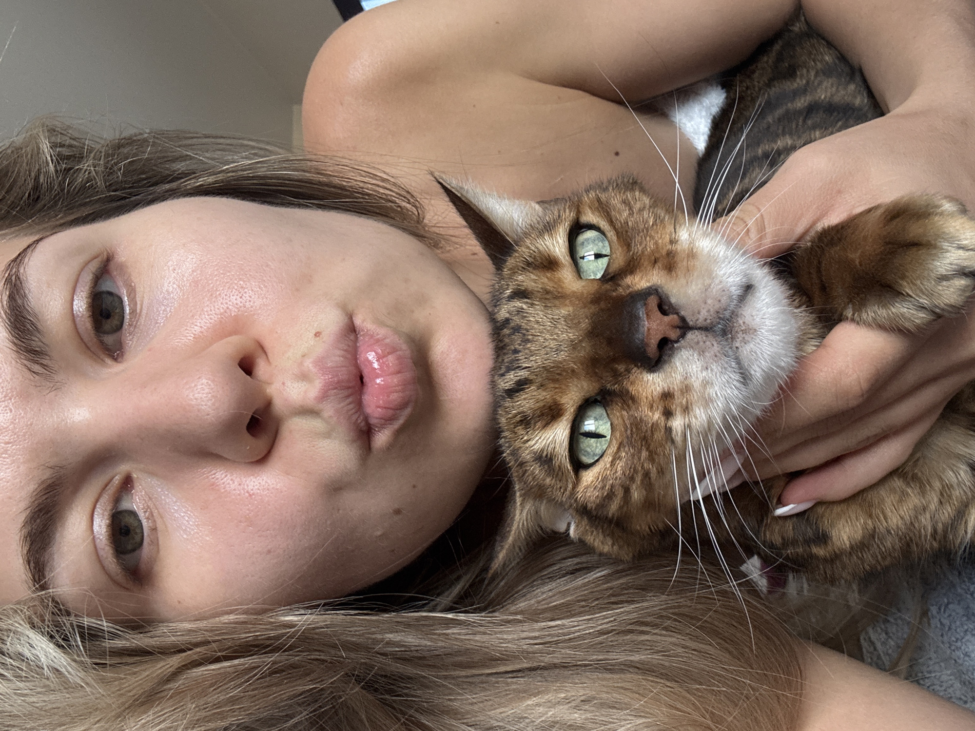 A medium close-up, head-and-shoulders shot shows a young woman with blonde hair, holding a brown and tan cat in her arms. The woman is facing the camera and puckering her lips, as if for a kiss. Her eyes are open and looking forward. The cat, which is positioned in front of the woman's face, has green eyes and is looking directly at the camera with a slightly annoyed expression. The woman's arms are visible around the cat, and her fingers are gently touching its fur. The background is out of focus, but appears to be an indoor setting. The lighting is bright, suggesting it is daytime.
