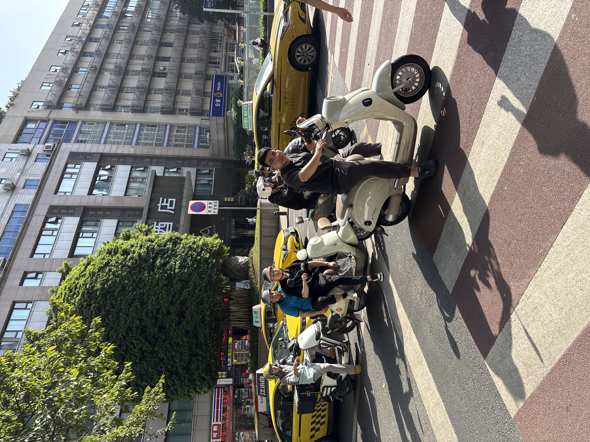 An outdoor, eye-level street view captures a bustling urban scene in Yuzhong District, China, under bright sunlight, indicated by the strong, long shadows cast across a distinctive beige and brown striped crosswalk. The image primarily features several individuals on white or cream-colored scooters/electric mopeds, seemingly paused at an intersection or in slow-moving traffic.
In the foreground, a man wearing glasses and a dark shirt sits on a scooter, looking directly at the camera. Behind him, another scooter carries three people, with the two visible passengers wearing helmets; one woman smiles broadly. Further down the street, another individual on a scooter is visible next to a yellow taxi. Several yellow taxis are parked or moving in the background, adding to the urban feel.
The background consists of tall, modern multistory buildings with numerous windows. One building prominently features green foliage growing on its exterior, while other trees line the street. A blue sign on a building reads "公安 POLICE," indicating a police station. Another sign partially visible near the street reads "凯旋路 严管路段" (Kaiyuan Road, Strictly Managed Section), and a red circular no-entry or no-stopping sign is also visible. Text on a taxi includes "监督电话 12345" (supervision hotline) and "军科整形焕新升级 5A形医院100整形专利" (military science plastic surgery upgrade, 5A hospital, 100 plastic surgery patents) is seen on a building. Other Chinese characters relating to a hotel, cigarettes, and alcohol brands are present on various signs. The overall impression is one of a busy city street on a clear, sunny day.