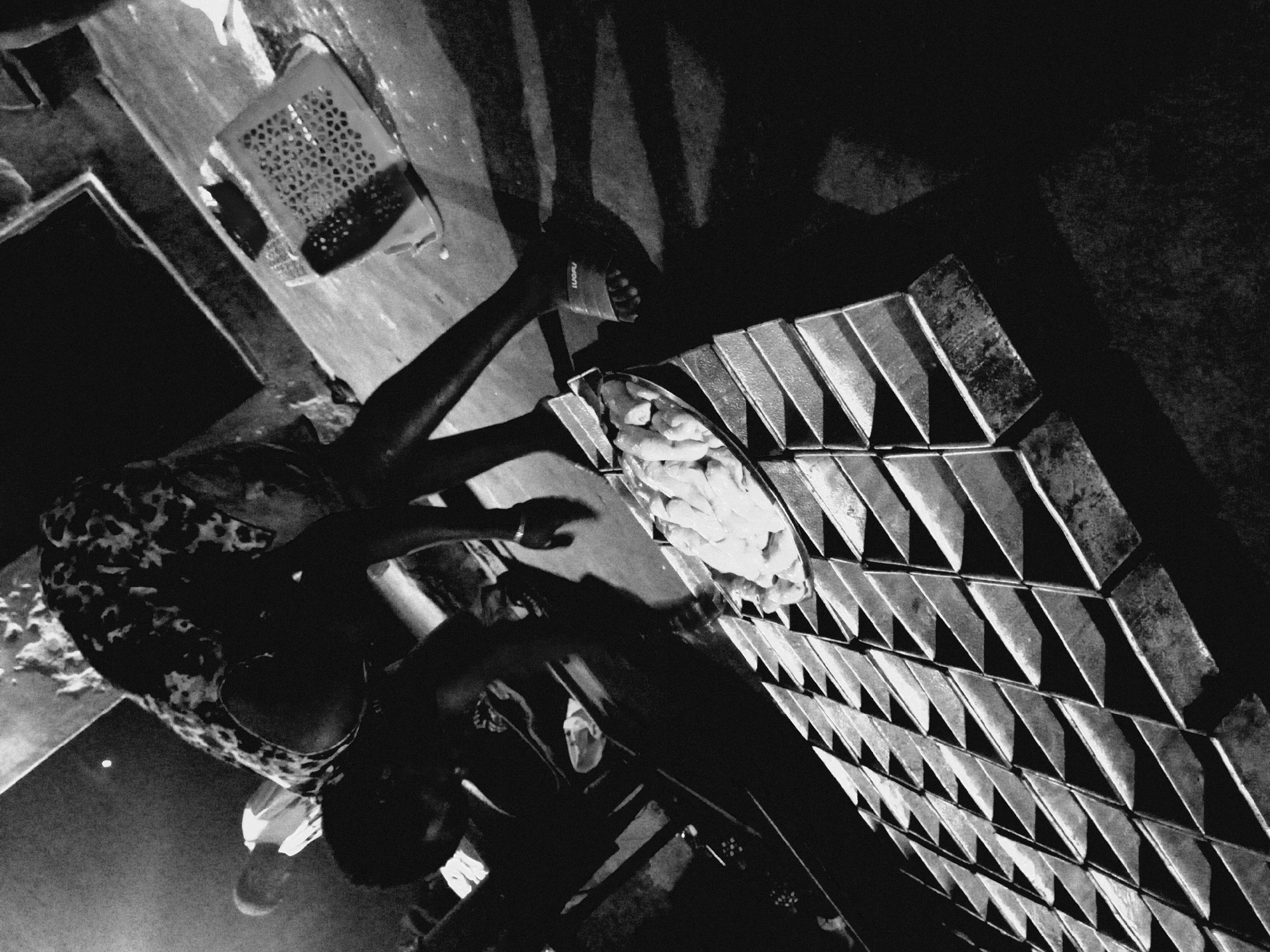 The high-contrast black and white photograph presents an angled, overhead perspective of a person engaged in a food preparation activity within a workspace. A figure, likely an adult, is visible on the left, wearing a patterned top and sandals. Their hands are positioned over a circular bowl filled with numerous light-colored, irregularly shaped food items, possibly raw dough or unbaked pastries.
To the right, a vast array of uniform, trapezoidal metal molds are meticulously arranged in tight rows, forming a strong, repeating geometric pattern across the surface. The person appears to be actively working with the contents of the bowl, likely transferring the food items into these molds, indicating a process related to baking or small-scale food production.
The scene is defined by dramatic, directional lighting, which casts long, sharp shadows across the floor and subjects. This stark contrast highlights textures and forms, plunging much of the background into deep shadow. In the upper left corner, a perforated plastic basket is faintly visible. The brand name "Melani" is discernible on the strap of a sandal worn by the person's foot. The image conveys a sense of focused, manual labor within a functional, utilitarian setting.