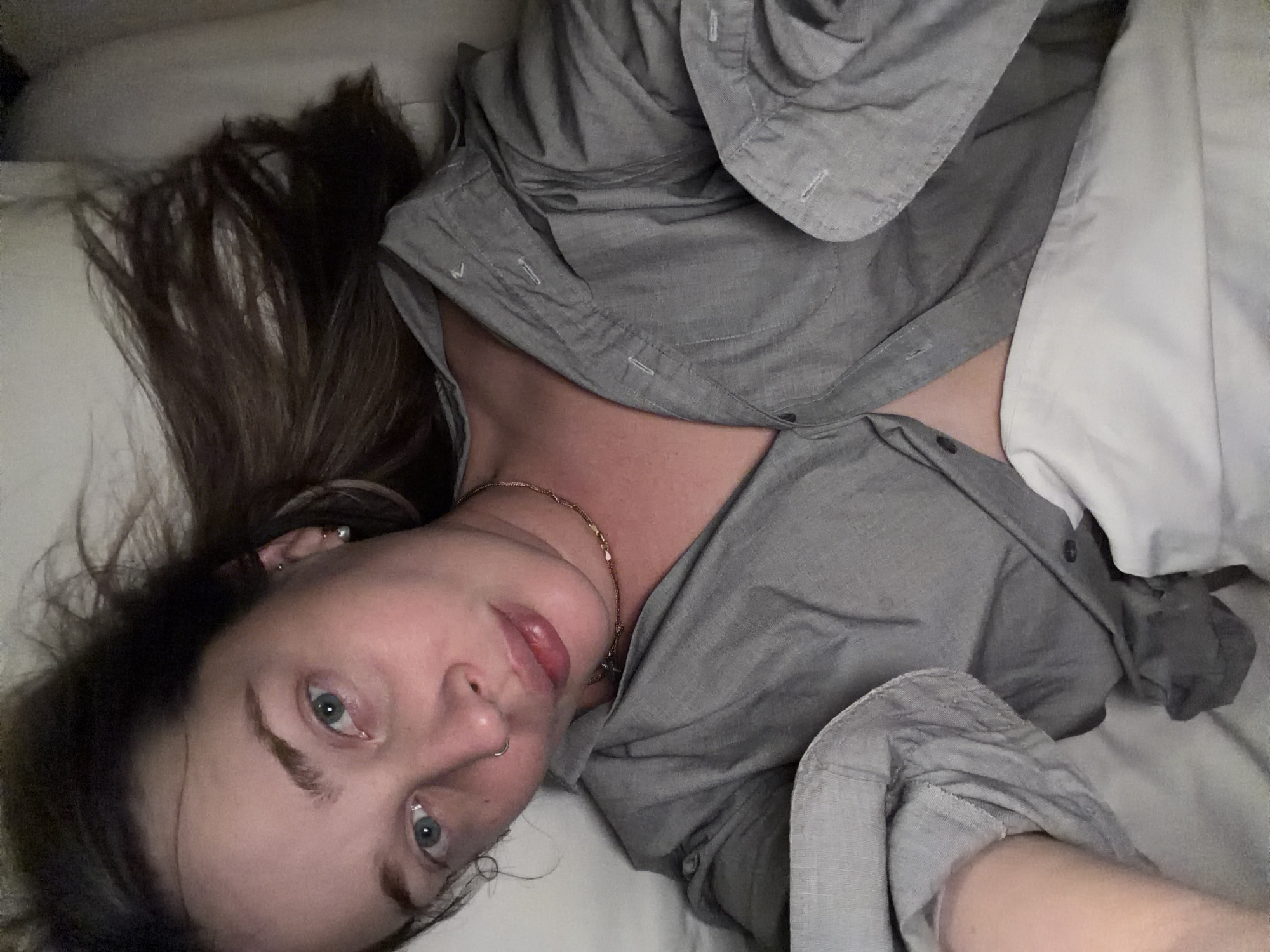 A woman lies on a white pillow, wearing a gray button-down shirt. Her hair is brown and disheveled. She has blue eyes, a nose ring, and a gold necklace. The location cannot be determined from the image.