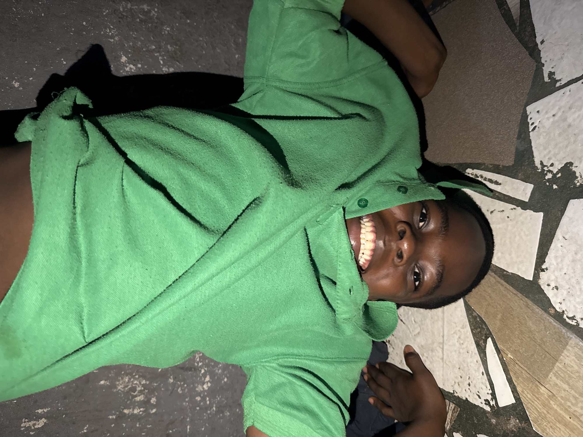 A young Black male is lying on his back, smiling broadly. He is wearing a green button-down shirt. He appears to be on a tile floor in what appears to be a residential area, in Onitsha, Nigeria. The floor has a patchwork pattern of various colors and shapes. There are shadows surrounding the male, and the overall lighting of the image is dim.
