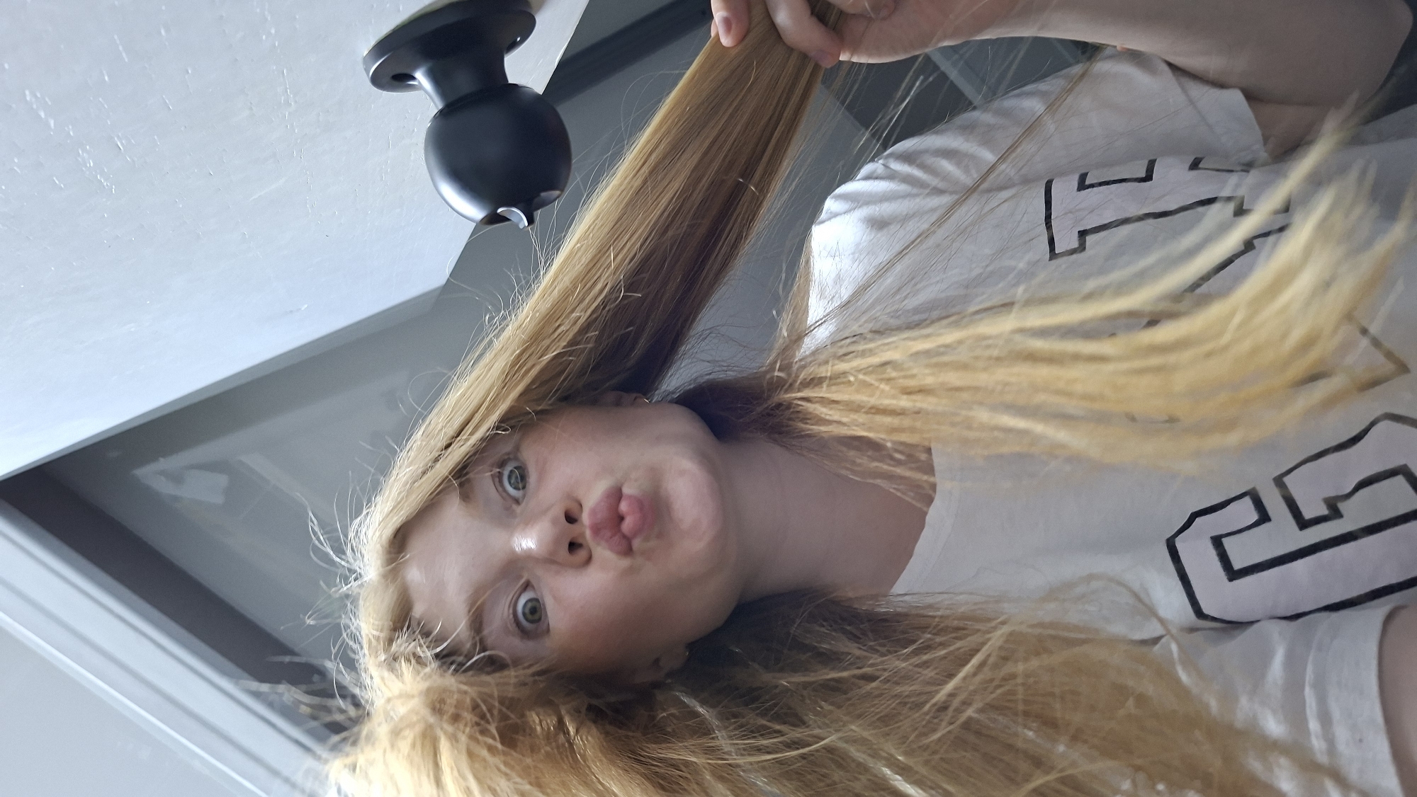 The image shows a close-up, low-angle, selfie-style shot of a young, fair-skinned woman with long, blonde hair that is messy and sticking out. She has light-colored eyes and freckles across her face. Her mouth is pursed in a kiss-like expression. She is wearing a white t-shirt with a large black "G" graphic on it. Her hair is being blown outwards, possibly by a fan or hair dryer, which is visible as a black object on the right side of the image, mounted on a wall. The background appears to be an interior space with light-colored walls and a corner where two walls meet. One of the walls has a grey, reflective surface. The overall lighting is bright and appears to be artificial. The scene does not contain any specific visual indicators that would allow for its identification as being located in the city of Chaikovsky, Russia.