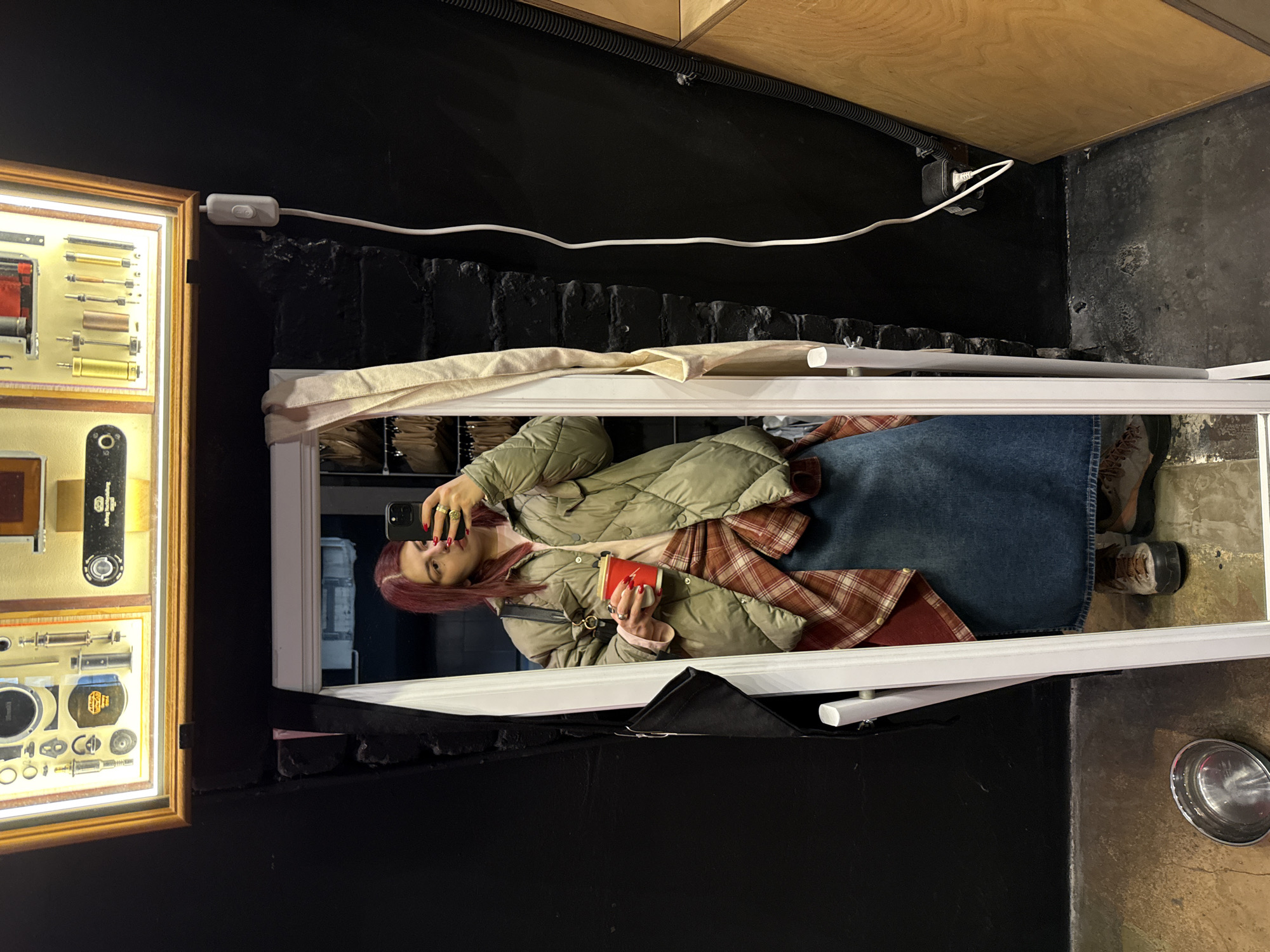 The image shows a woman taking a selfie in a mirror. She has reddish-purple hair and is wearing a puffy green jacket, a plaid shirt, and a denim skirt. She holds a red cup in one hand and her phone in the other. The mirror frame is white. To the left of the mirror is a framed display containing various tools and hardware. The background is dark, with a wooden ceiling and a black wall. There is a white electrical cord running along the wall and a light switch. A metal bowl is visible on the ground to the right of the mirror. I cannot identify if this is in Moscow, Russia, as there is no verifiable location.