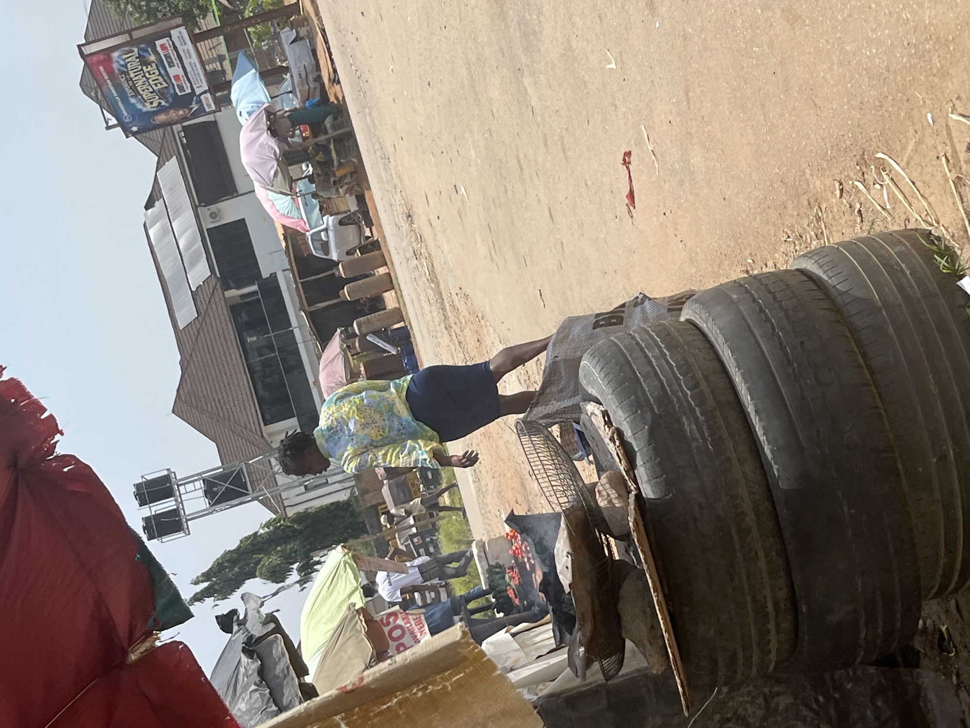 The image depicts an outdoor market scene in Abuja, Nigeria, under a bright, hazy sky. In the foreground, a stack of used tires is visible, partially obscuring a makeshift cooking station. This station features a metal grate over burning coals, with raw tomatoes and other vegetables arranged on top, suggesting food preparation.
A woman in a vibrant, floral-patterned shirt and a dark skirt is the central human figure, walking away from the camera towards the market stalls. She appears to be a vendor or a shopper. Behind her, other market activities are taking place. Several umbrellas of varying colors provide shade over stalls where goods are displayed. A white SUV is parked nearby, and in the background, a multi-story building with a prominent billboard for "SUPERDRAGON EDGE" is visible. The environment is dusty, with a dirt road running through the market area. A water tower stands tall on a metal structure, adding to the urban infrastructure visible in the scene. The overall impression is one of a busy, open-air marketplace.