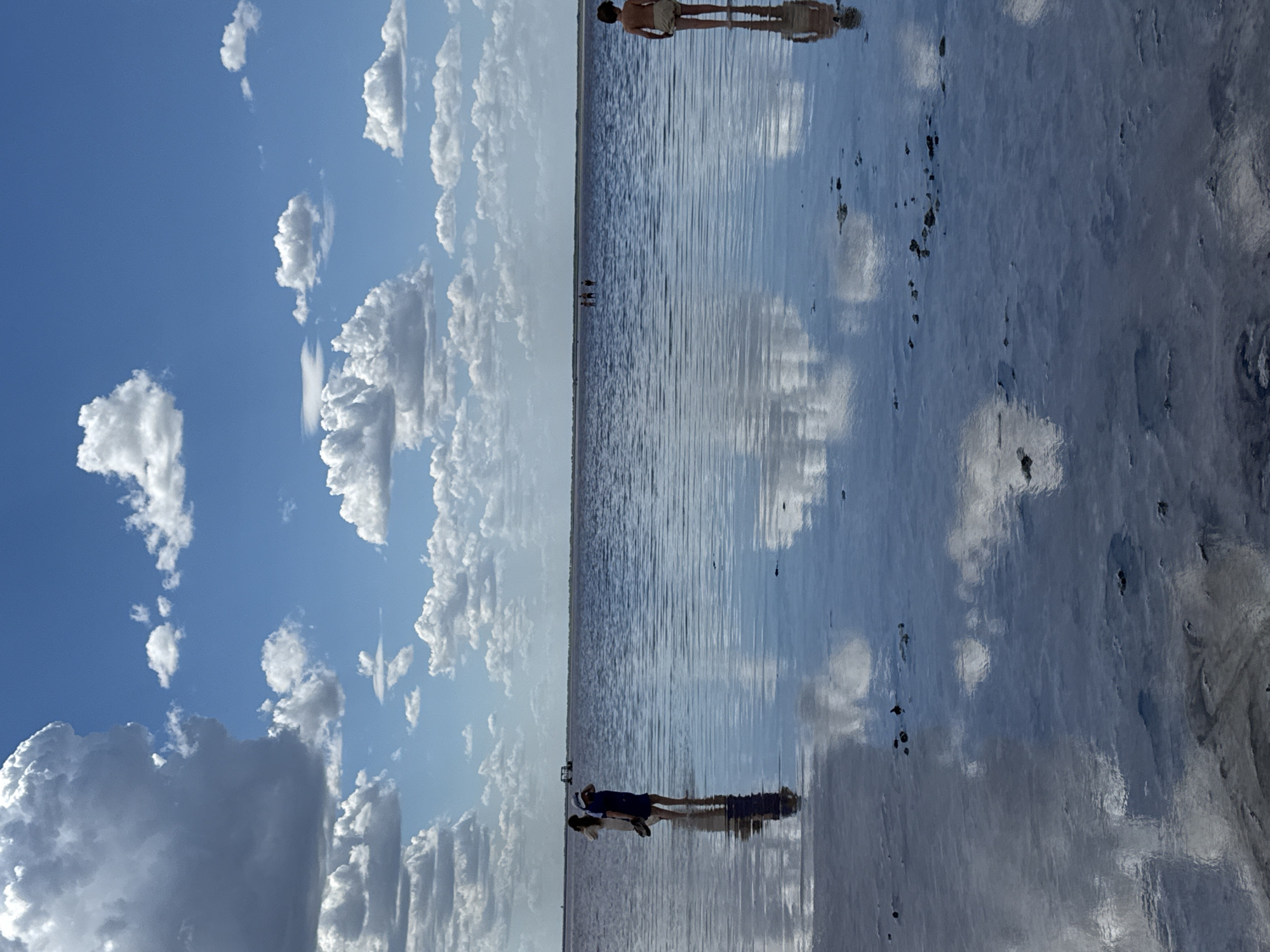 The image depicts a shallow body of water, likely a salt lake, reflecting the sky and clouds. Several individuals are visible wading in the water. On the left, two people stand close together, one appearing to be taking a photo. Further in the distance, a group of people are scattered across the water. On the right side of the frame, a single child stands facing away from the camera. The environment is open with a clear blue sky dotted with fluffy white clouds. The water is calm, with ripples distorting the reflections of the sky and the figures. The location is identified as Burbol, Russia.