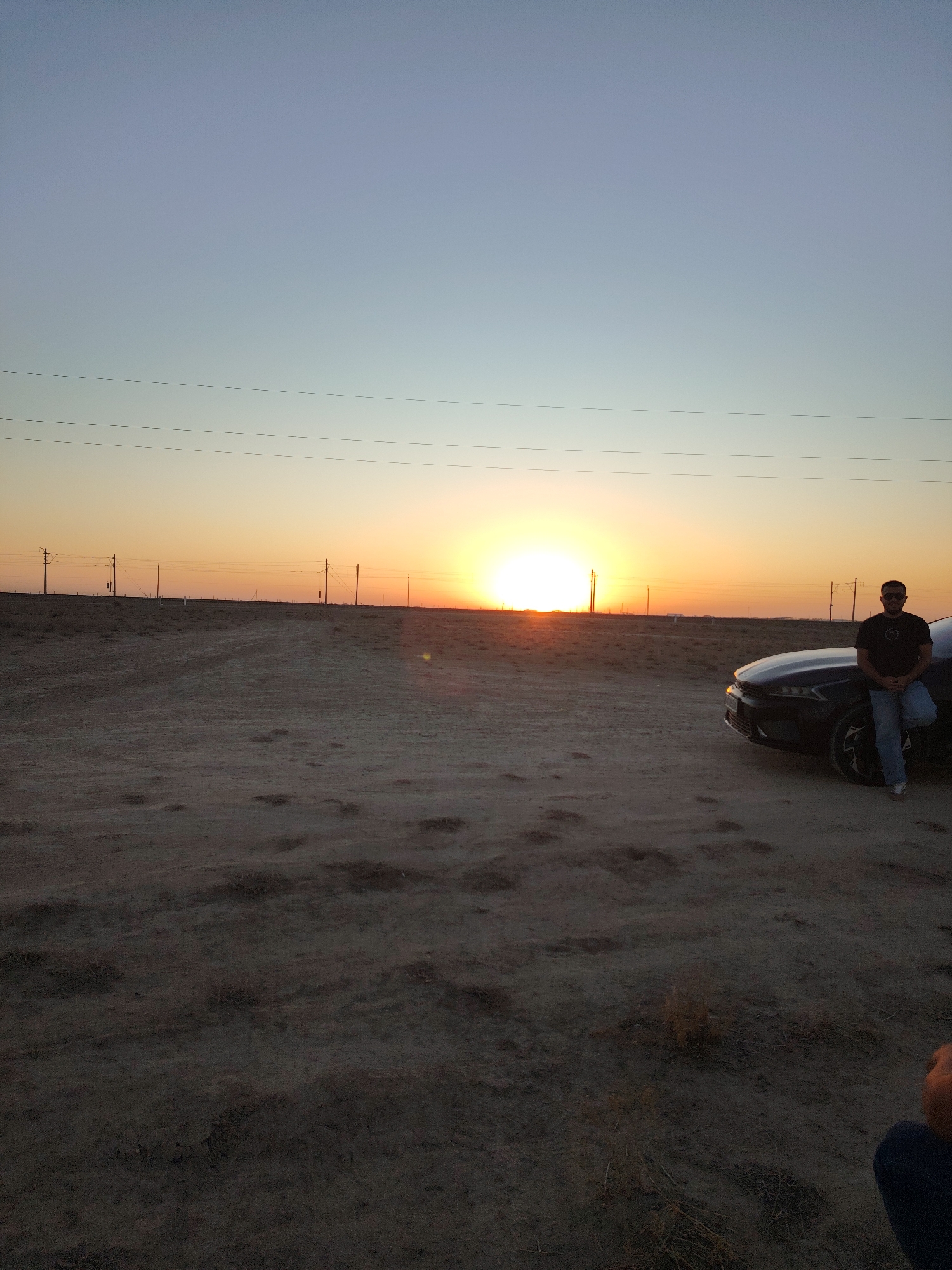 In Haratan, Uzbekistan, at sunset, a man in a black t-shirt, blue jeans, and sunglasses leans against a dark-colored car parked on a dirt road. The sun is setting on the horizon, casting a warm glow across the sky, which transitions from a pale blue at the top to a vibrant orange and yellow near the horizon. Power lines stretch across the background, supported by poles. The terrain is dry and dusty, with sparse vegetation.