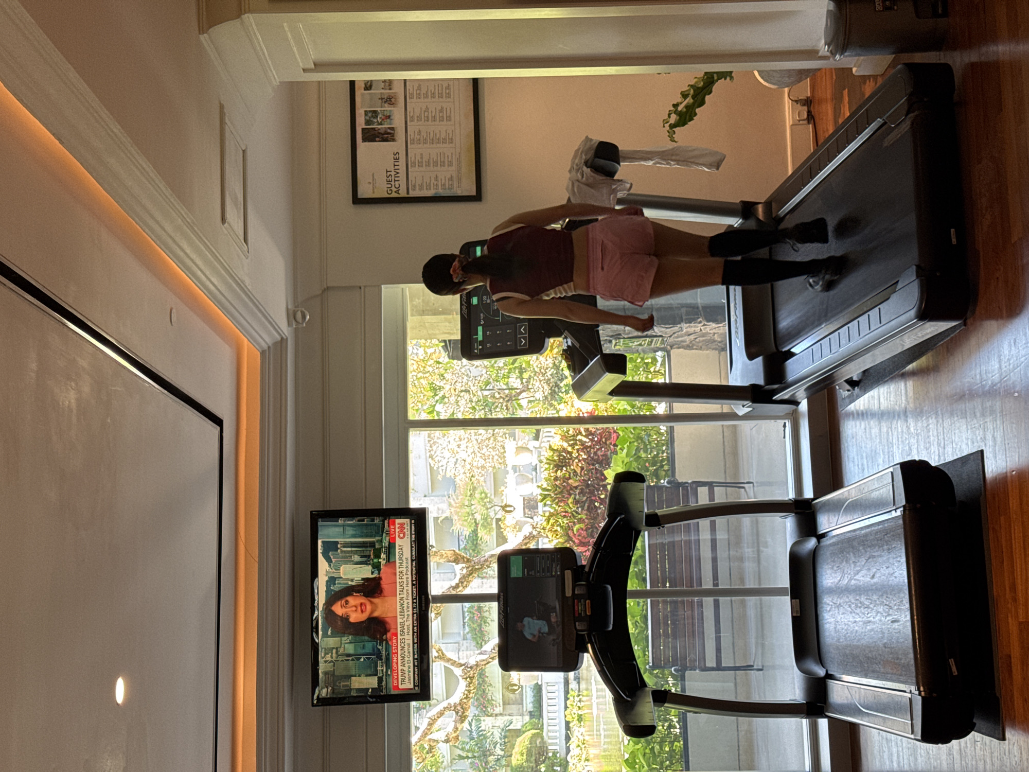 A woman with dark hair in a red and white tank top and pink shorts walks on a LifeFitness treadmill inside a room. A CNN broadcast with Jasmine El-Gamal appears on a television. Another treadmill sits nearby. A window looks out onto trees and a bench. A list of "GUEST ACTIVITIES" hangs on the wall.