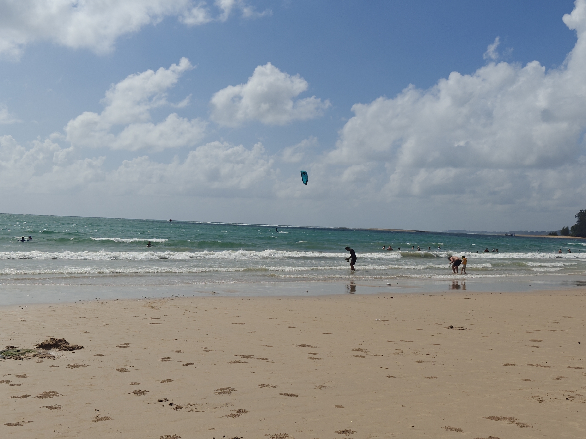 The image depicts a wide, sandy beach in Sakhu, Thailand, under a partly cloudy blue sky. The turquoise ocean stretches to the horizon, with gentle waves breaking on the shore. In the distance, a teal kiteboard is visible in the air, suggesting the presence of kitesurfing activities. Several people are in the water, some swimming and others appearing to be engaged in watersports. Closer to the shore, two individuals are standing in the shallow water, one a darker-skinned adult in dark shorts and the other a lighter-skinned child in a yellow shirt. The foreground of the image is dominated by the sandy beach, with small mounds of sand and some debris scattered across it. The overall scene conveys a leisurely day at the beach, with a mix of recreational activities.