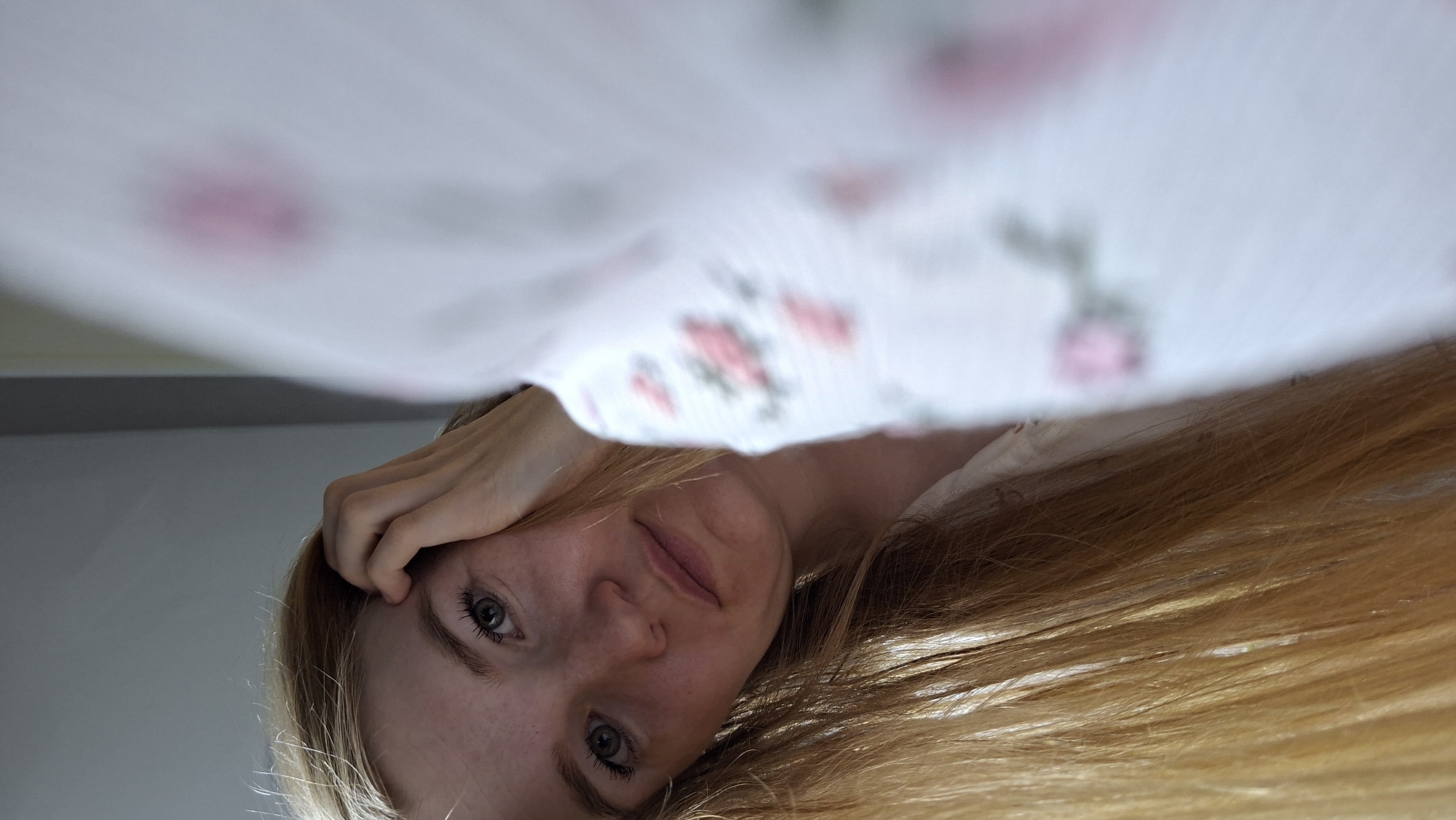 A young woman with long blonde hair rests her head on her hand, looking towards the camera. She has green eyes and freckles across her nose and cheeks. She is wearing a white top with a floral pattern. The background is a plain grey wall. The image appears to be taken indoors, possibly in a bedroom or dressing area. There is no indication of the location in the city of Chaikovsky, Russia.