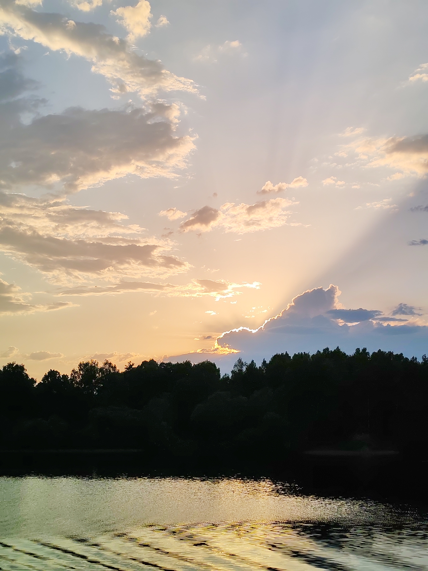 The image depicts a sunset scene, likely taken at dusk, in Krasnaya Gorka, Russia. The primary focus is on the sky, which features a mix of clouds in various shades of white, gray, and pale orange, illuminated by the setting sun. The sun's rays stream downward, partially obscured by a prominent cloud formation in the center of the frame. The light is golden, suggesting the sun is low on the horizon.
Below the sky, a dark, silhouetted treeline stretches across the middle ground. The trees appear to be dense and form a dark barrier between the sky and the water below.
In the foreground, a body of water reflects the colors of the sky and the setting sun. The water surface has subtle ripples or waves, visible through the light reflecting off of it.
There are no people visible in the image. The overall scene conveys a sense of tranquility and natural beauty.