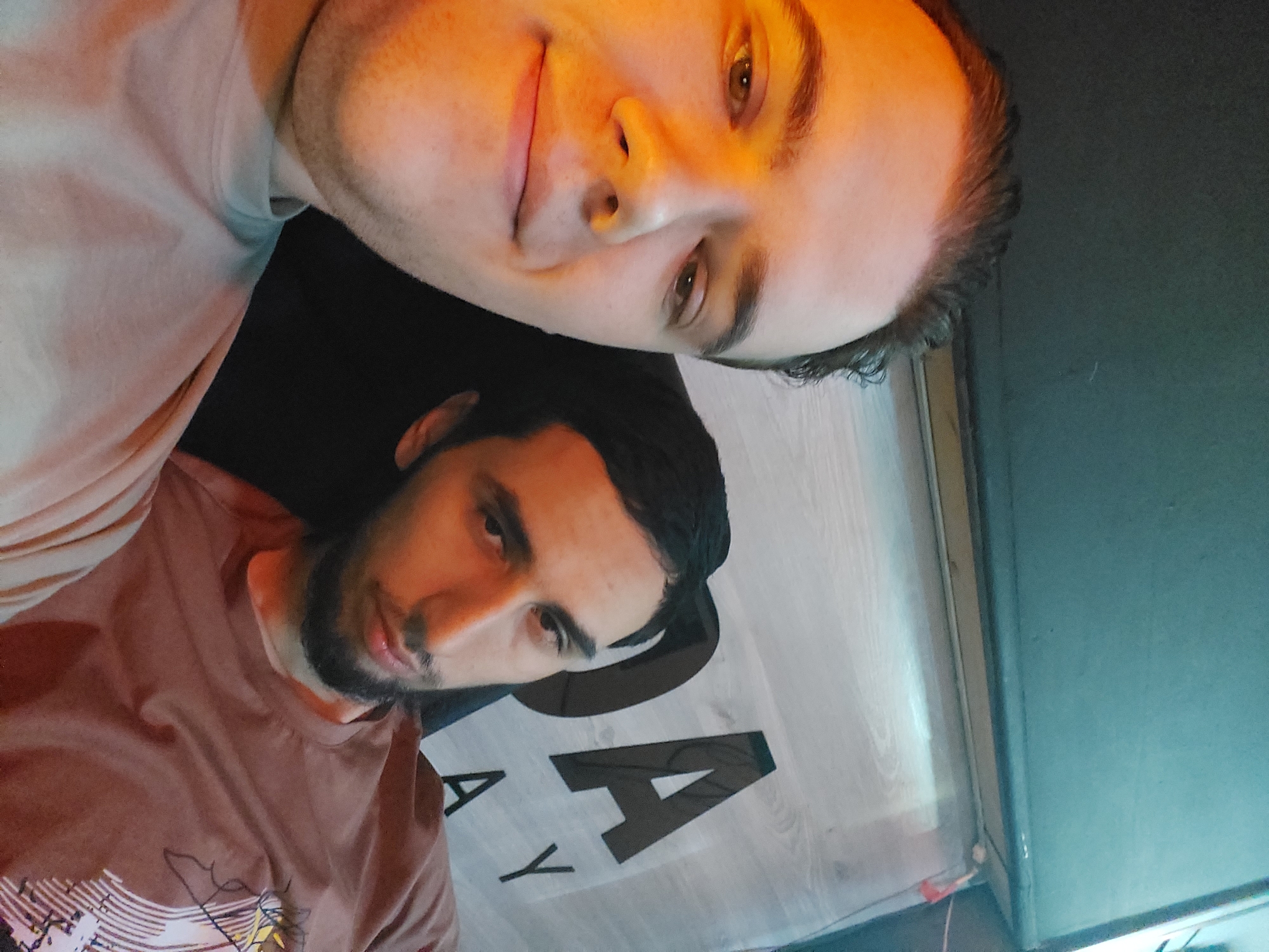 Two men are visible in the image. The man at the top of the image is fair-skinned, has short dark hair, and is wearing a light-colored t-shirt. He is looking at the camera and has a slight smile. The man at the bottom of the image has darker skin, a dark beard, and is wearing a brown t-shirt. He is also looking at the camera. Behind them is a light-colored wall with the partially visible letters "DAY". The background appears to be indoors. The location may be Tashkent, Uzbekistan. Lighting appears to be artificial and somewhat warm, possibly from an overhead light.