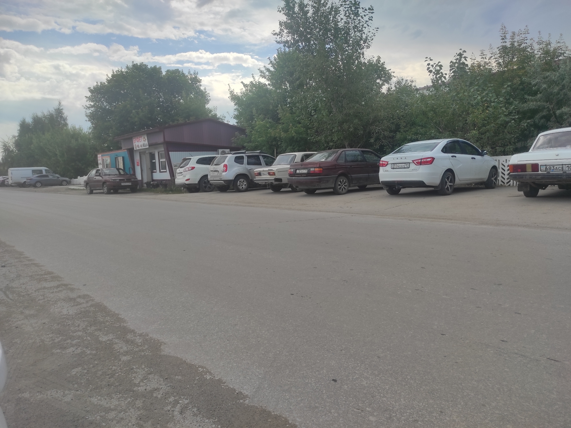 This image shows a row of parked cars on the side of a paved road in front of a small building with a maroon roof. The building appears to be a small shop or kiosk with signs in Cyrillic script. The cars are various models and colors, including a white sedan with a license plate that reads "E554XH 10". Trees and foliage line the background, and the sky is cloudy. The scene is likely located in Denisovka, Kazakhstan, given the Cyrillic script and the general appearance of the environment. There are no people visible in the image.