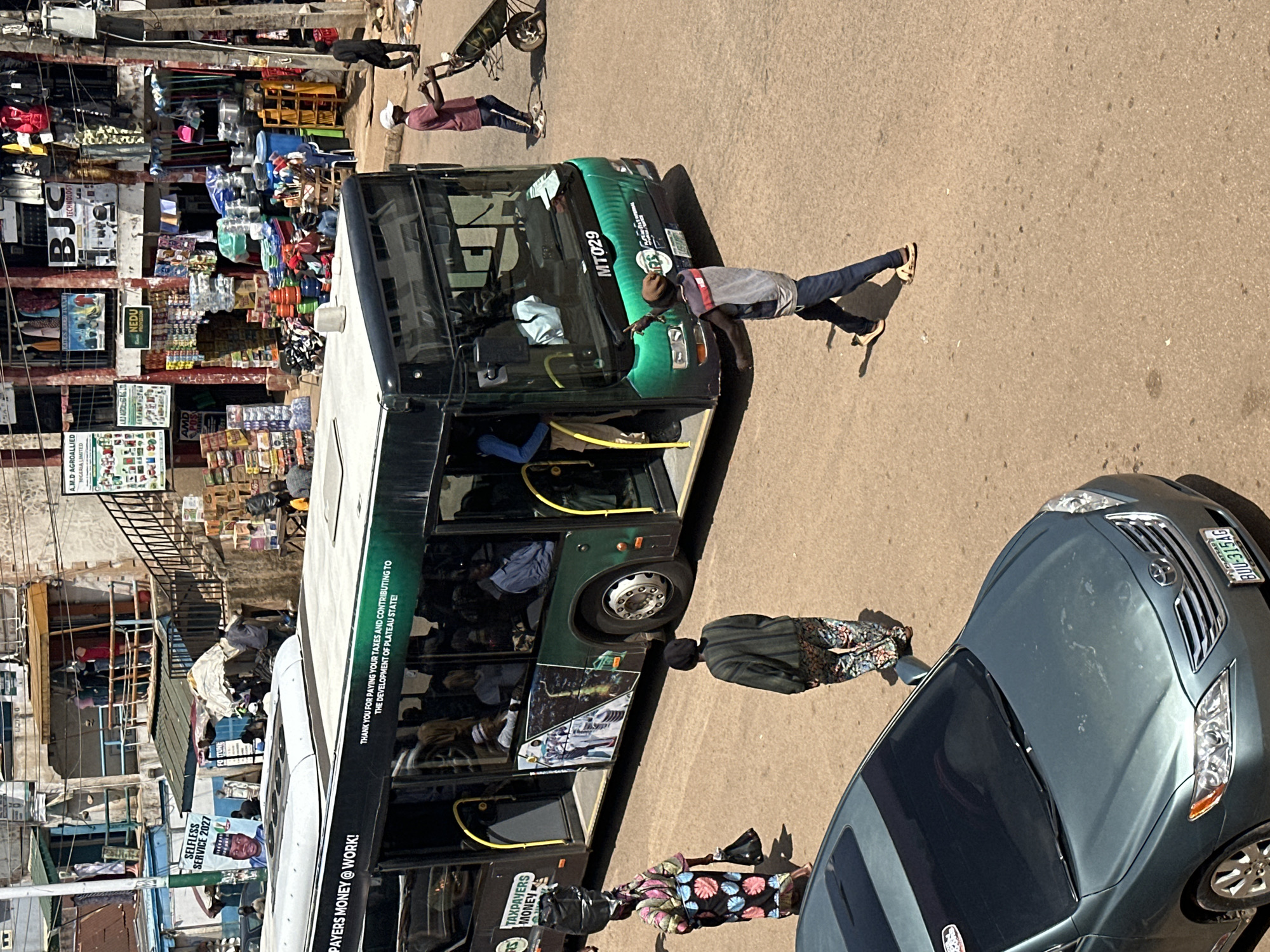 In the city of Bukuru, Nigeria, a street scene unfolds with a focus on a green and white bus. The bus, labeled "MT029" and bearing the inscription "TAXPAYERS MONEY @ WORK! THANK YOU FOR PAYING YOUR TAXES AND CONTRIBUTING TO THE DEVELOPMENT OF PLATEAU STATE!", is positioned centrally. Its doors are open, revealing passengers inside.
To the left of the bus, a woman in a vibrant, patterned dress and headwrap walks away from the viewer, carrying two dark bags. A second woman, in a dark top and patterned trousers, walks towards the right of the frame, her back to the camera.
In the foreground, a dark grey sedan is parked. Its license plate reads "BUU-315AG". Further to the right and slightly behind the bus, a man in a maroon shirt and dark pants pushes a wheelbarrow. Another man, wearing a light grey shirt, dark pants, and a brown beanie, walks towards the foreground, his left arm raised. He has a reflective vest or bag slung over his shoulder.
The background consists of multi-story buildings lining the street, with shops displaying various goods, including clothing, kitchenware, and packaged items. Some of these shopfronts have signage, with visible text such as "BJC TECHNOLOGY" and advertisements. The overall environment appears to be a busy urban market area.