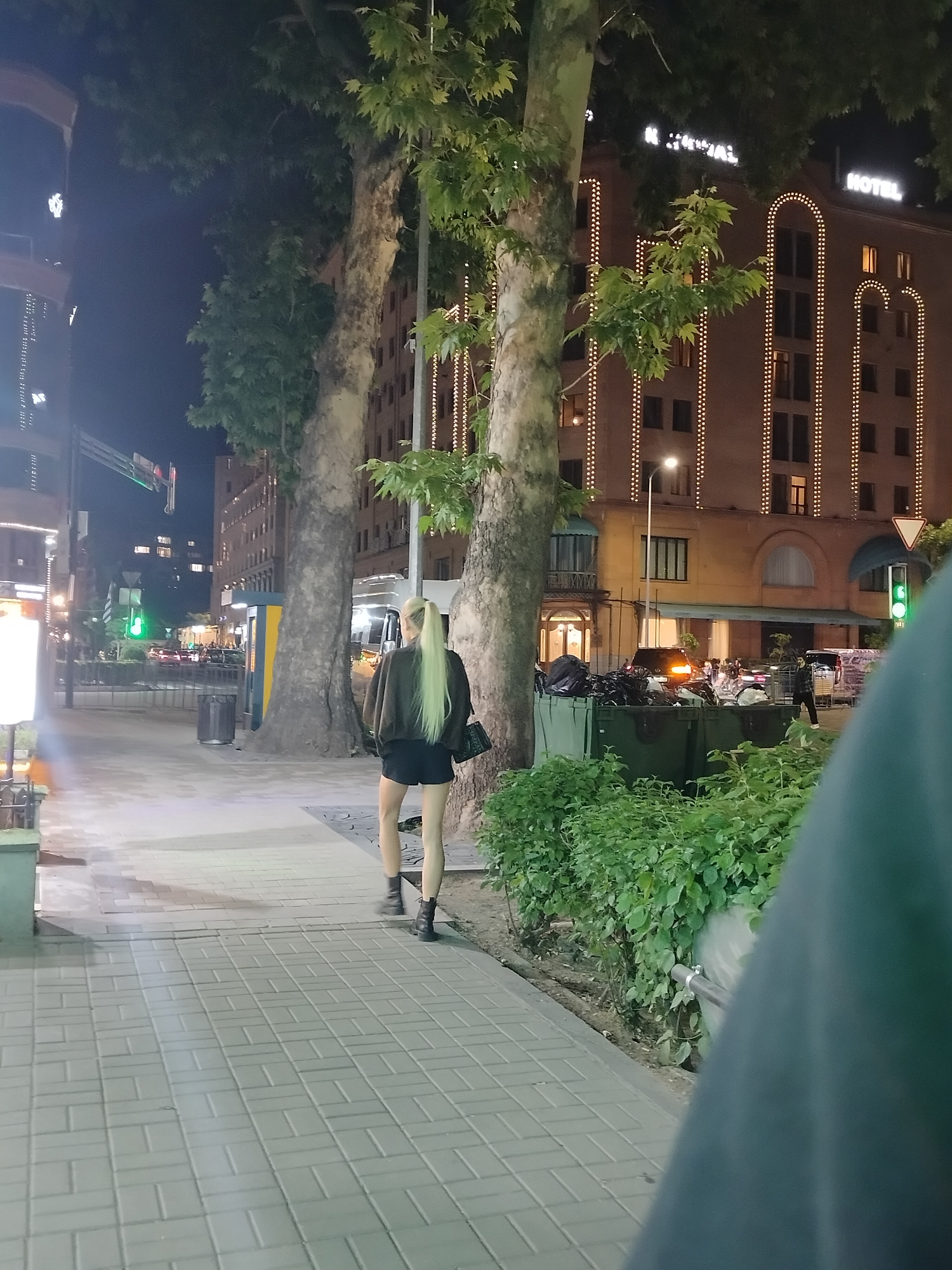 The image shows a nighttime scene on a sidewalk in Yerevan, Armenia. A woman with light-colored, long hair in a ponytail is walking away from the camera. She is wearing a dark jacket, dark shorts, and dark boots, and carrying a dark bag.
Behind her, two large trees flank a yellow and blue street booth. In the background is a multi-story building identified as the "National Hotel" with lights adorning its facade. There is street lighting, a traffic signal, and other urban features like trash bins and bushes. Cars and people are visible further down the street. The sidewalk is paved with rectangular blocks. A person is partially obscuring the foreground on the right side of the image.