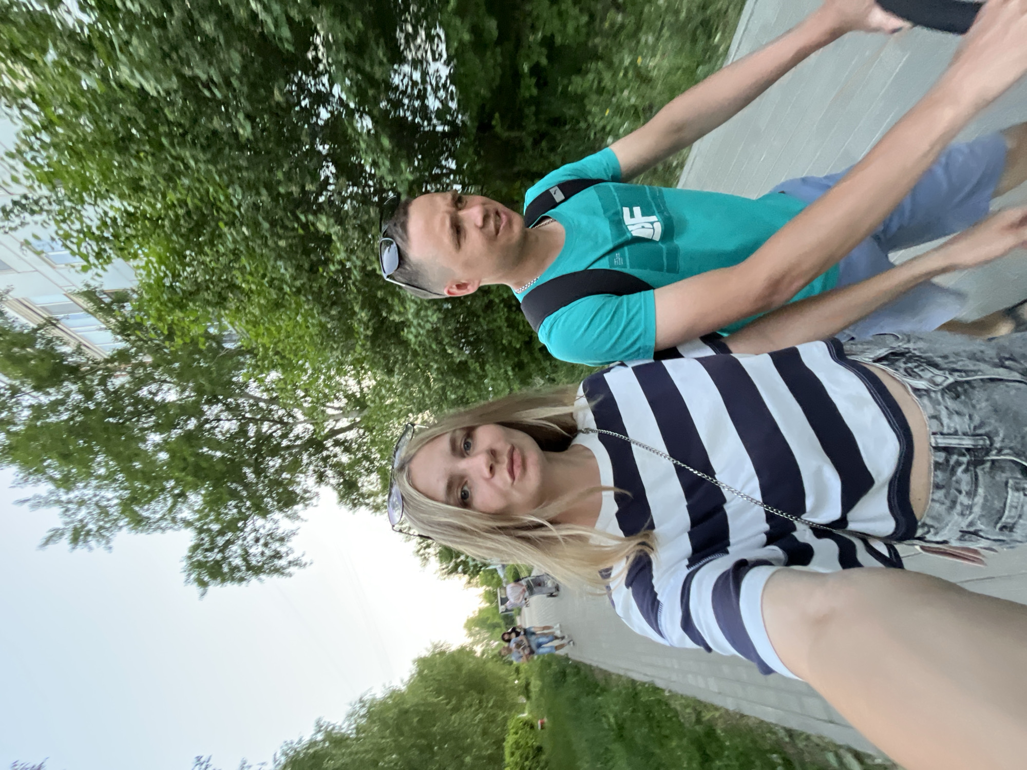 Two individuals are pictured in what appears to be a selfie taken along a walkway. The woman, positioned in the foreground, wears a striped shirt and has blonde hair. Behind her is a man with a teal shirt and sunglasses perched atop his head. Both individuals seem to be in motion. The walkway is situated in a green, outdoor environment, likely a park, with trees visible on either side. Other people are seen walking further down the path. Based on the camera angle and the presence of modern infrastructure, it is plausible that the location is in the city of Chaikovsky, Russia, given that it is one of the possible locations, though definitive verification would require more information about the specific surroundings.