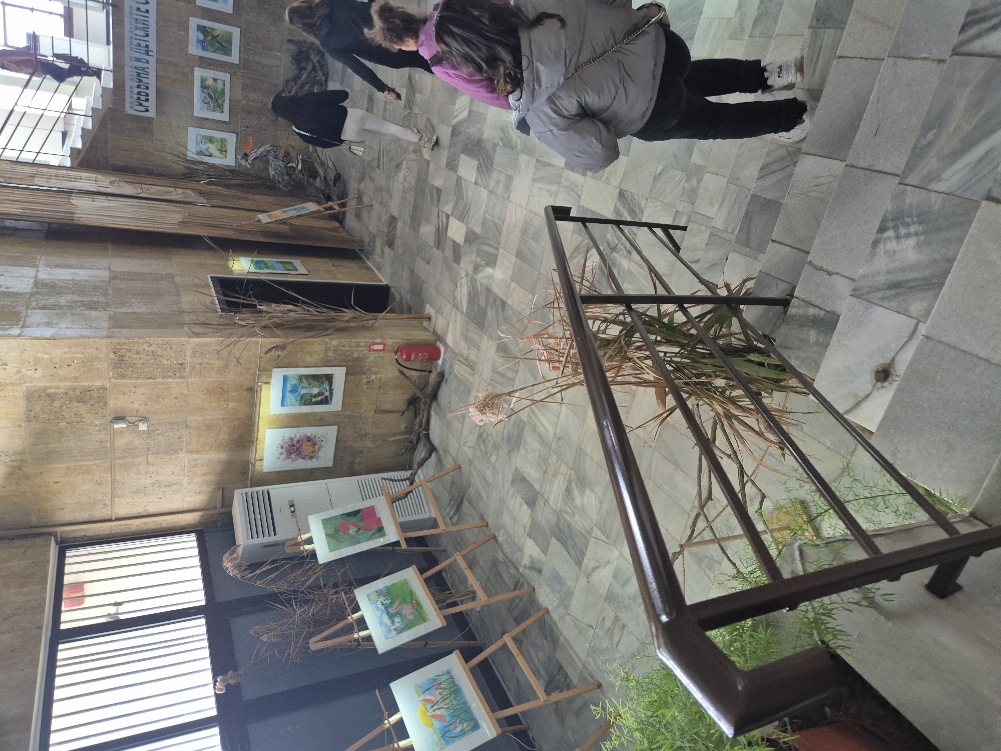 The image shows an indoor exhibition space in Srebarna, Bulgaria. Several paintings are displayed on easels and mounted on the wall, featuring themes such as landscapes and animals. There are visitors viewing the exhibition. The space has a tiled floor, and a staircase with a metal railing is present. A fire extinguisher is mounted on the wall. There are dried floral arrangements and decorative elements. The architecture appears to be a combination of exposed stone or concrete walls and large windows with blinds.