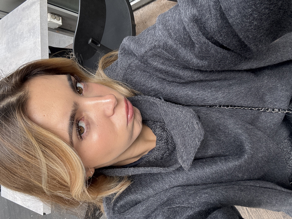 The image depicts a close-up, chest-up selfie of a single female subject, approximately 20-35 years old, in the city of Vaulx-en-Velin, France. She has light skin, long brown hair with prominent blonde highlights, parted centrally, and brown eyes looking directly into the camera. Her lips are glossy and positioned in a deliberate pout. A small, light-colored stud earring is visible in her right earlobe. She is wearing a dark grey, textured overcoat or jacket with an unstructured, possibly cowl-neck collar, layered over a darker grey, possibly knitted, garment. A thin, dark-colored chain, likely from a bag, is visible crossing her chest.
The background, partially visible on the right, shows a section of a light grey, rectangular table surface. Below the table, a dark-colored chair with a rectangular cutout in its backrest is present. An off-white electrical outlet or fixture is affixed to a vertical surface adjacent to the table. A light brown, textured floor or ground surface is visible at the bottom right. A lighter, possibly white, vertical surface is faintly discernible in the upper left background. The overall scene suggests an indoor or sheltered outdoor setting.