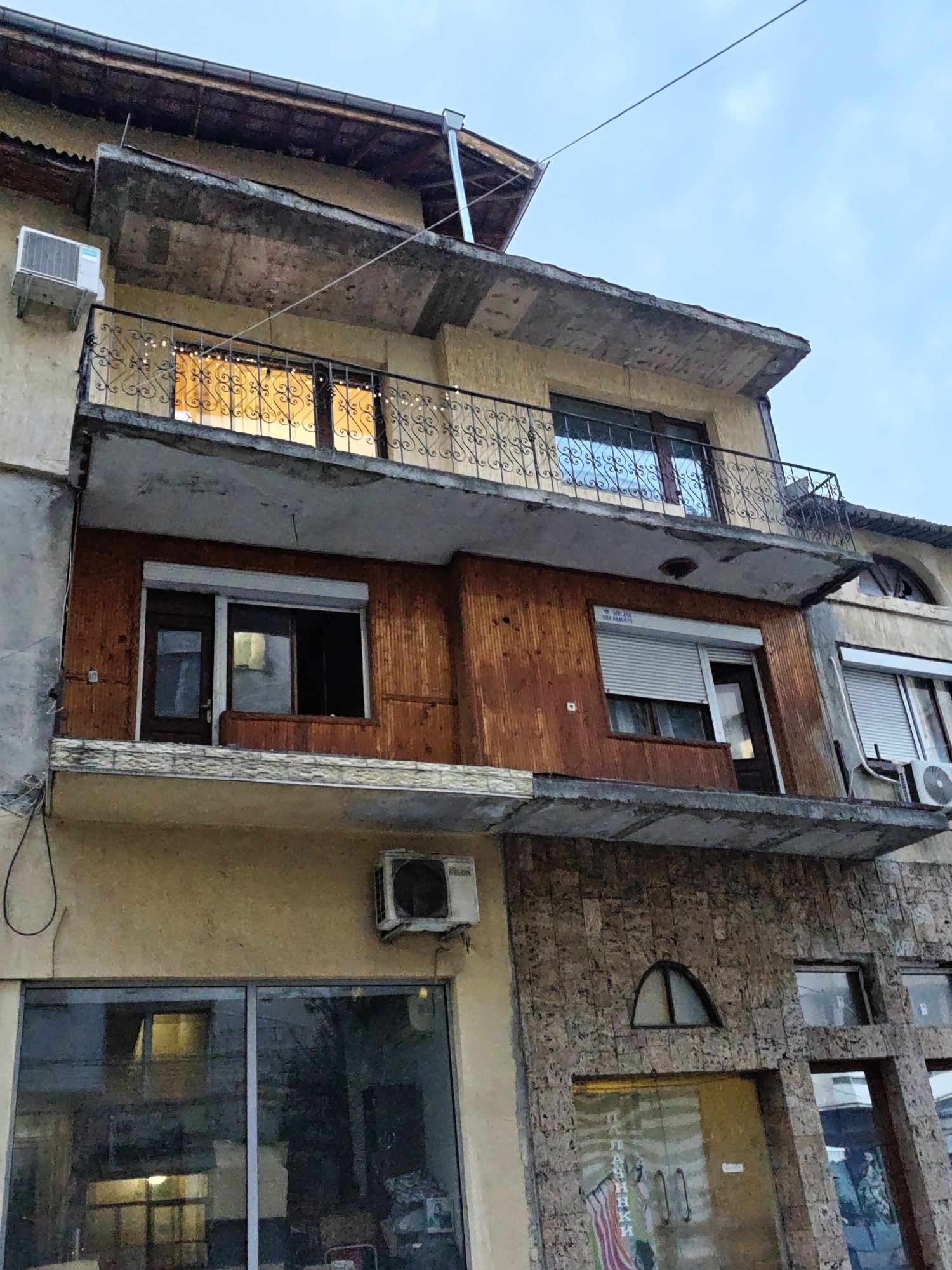 The image depicts the facade of a multi-story building in the city of Lovech, Bulgaria. The building features balconies with decorative metal railings on the upper floors. One balcony has large windows with warm light emanating from within, suggesting interior lighting. Below this, another section of the building is clad in dark wood paneling and has windows, some with closed rolling shutters and one with an open shutter revealing a dimly lit interior. There are also air conditioning units mounted on the exterior walls. The lower part of the building is faced with stone and features a large glass storefront with reflections of the surroundings. The sky above is overcast with a pale blue hue.